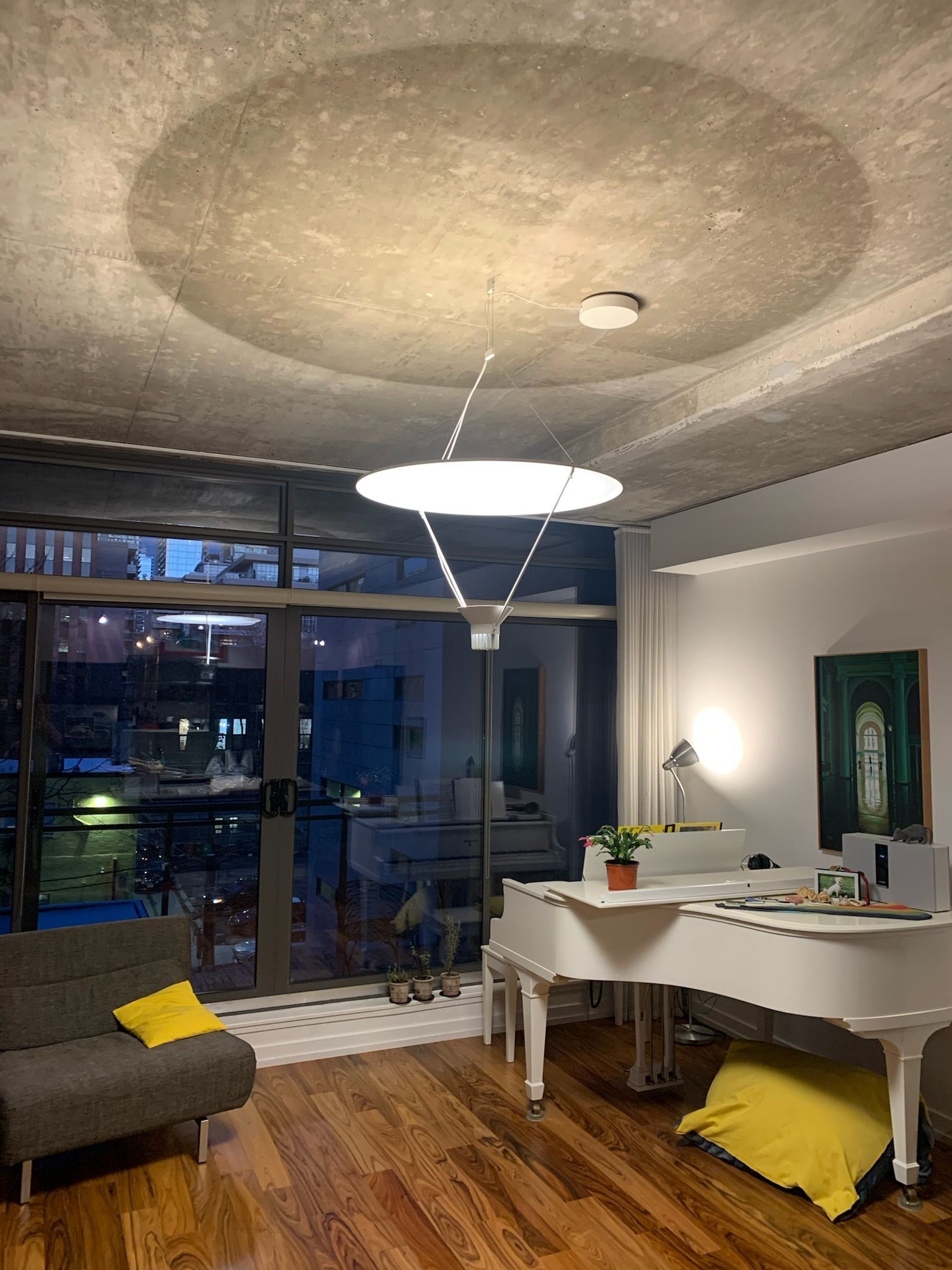Modern living room with a white piano, dark gray sofa, and a large window overlooking a city at dusk.