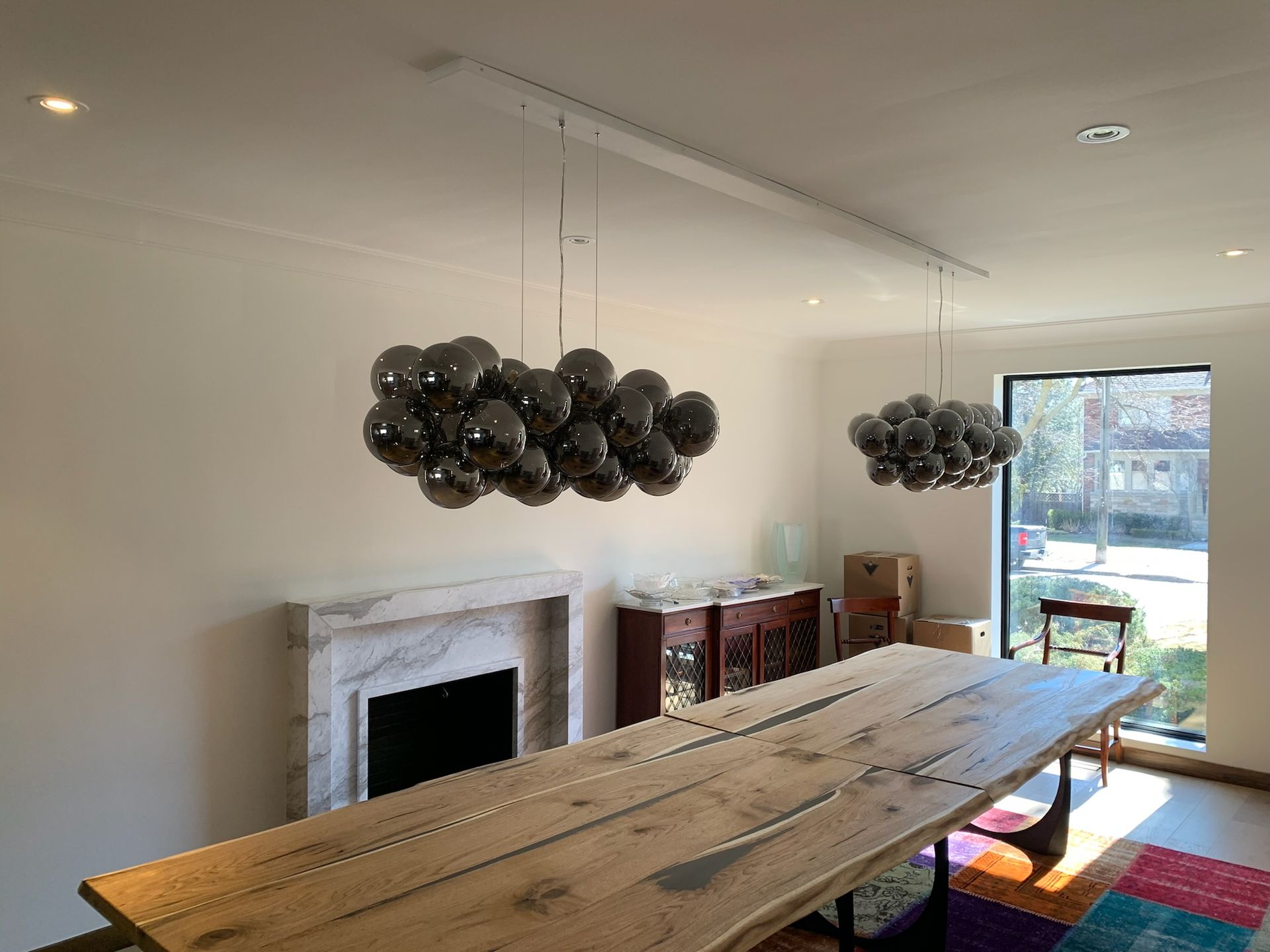 Long wooden dining table with two clustered pendant lights, fireplace, and window.