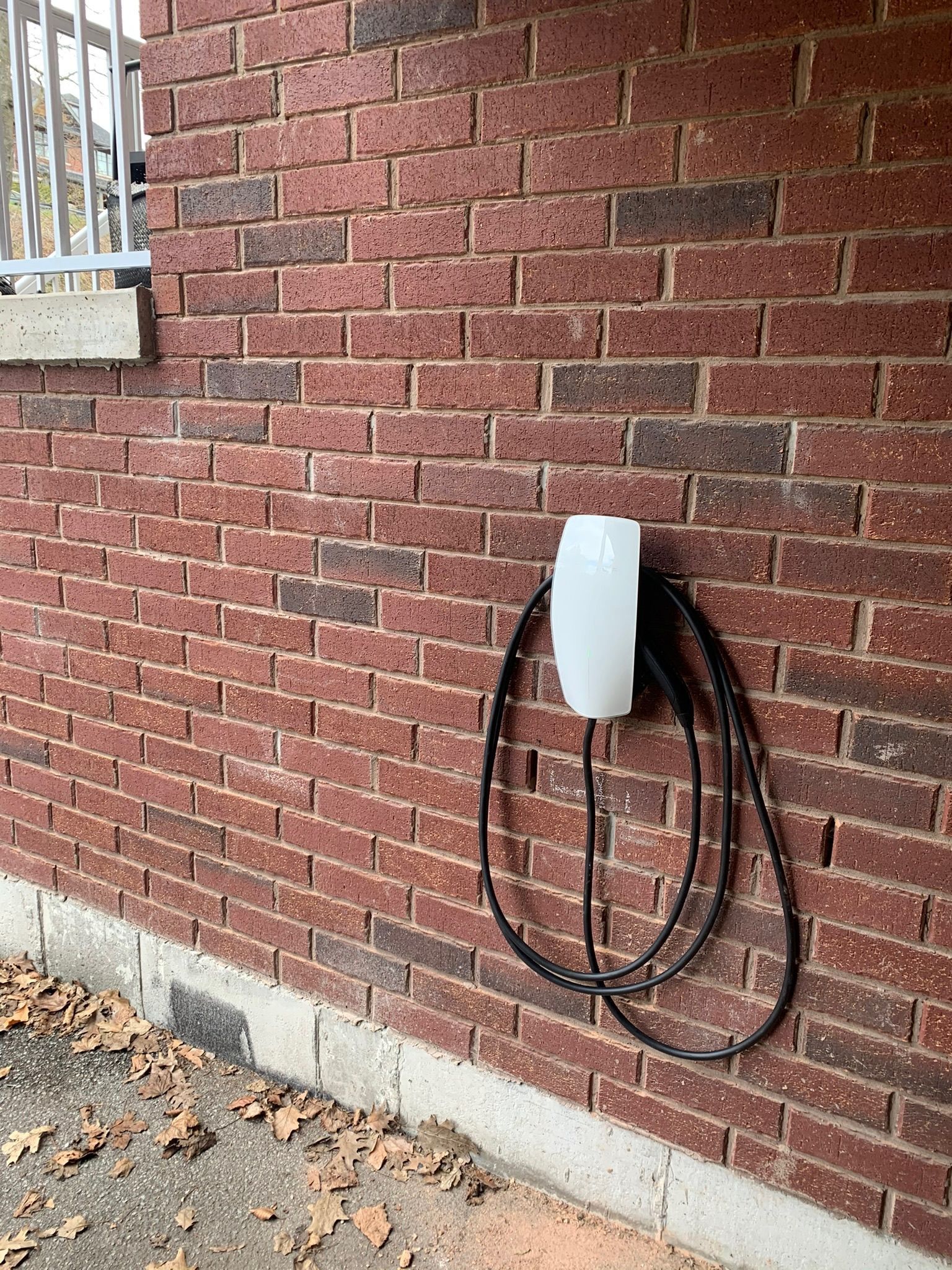 A white electric vehicle charger is mounted on a red brick wall. The charging cable is looped below.