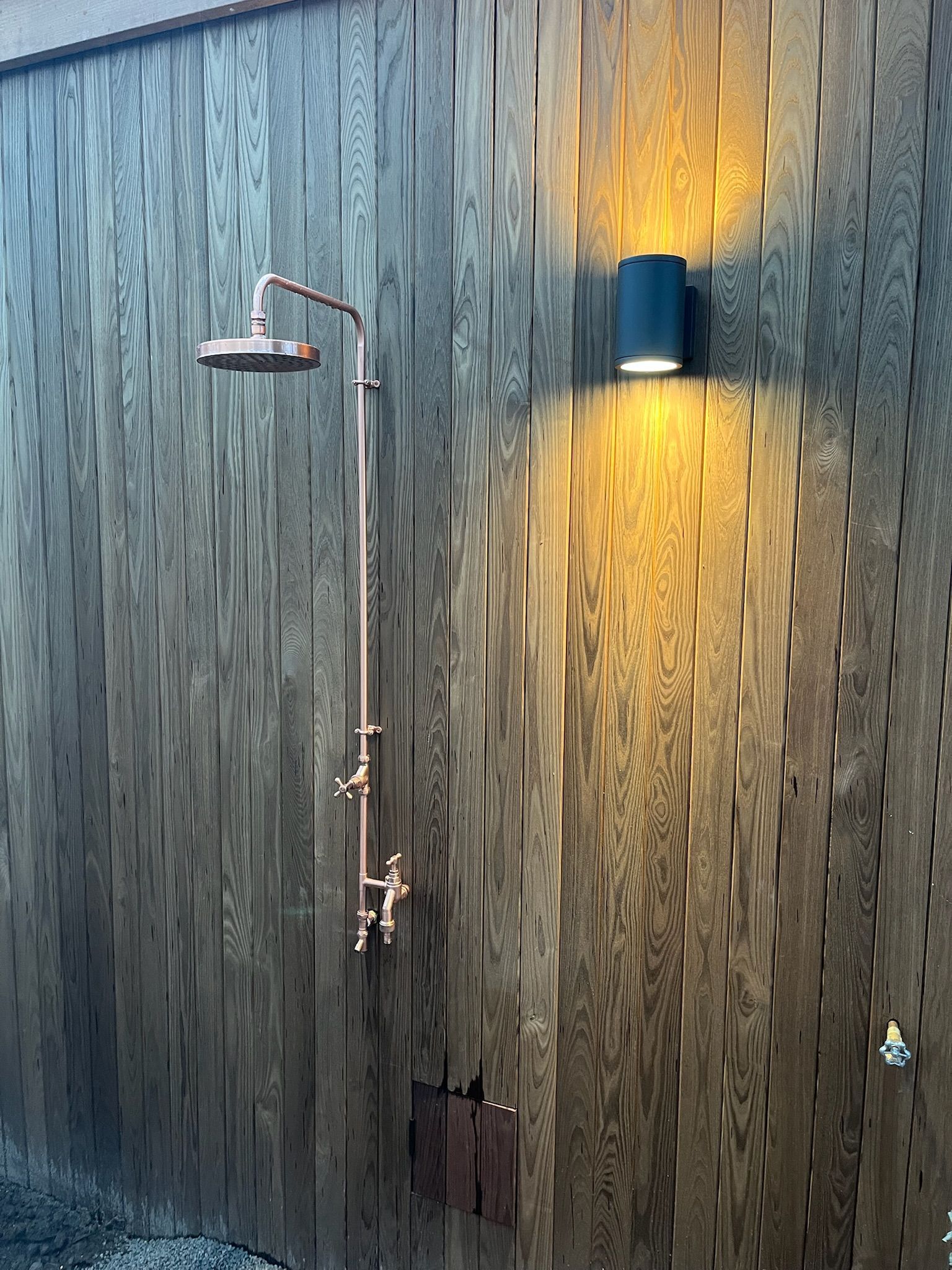 Outdoor copper shower against weathered wooden wall, with light fixture above.