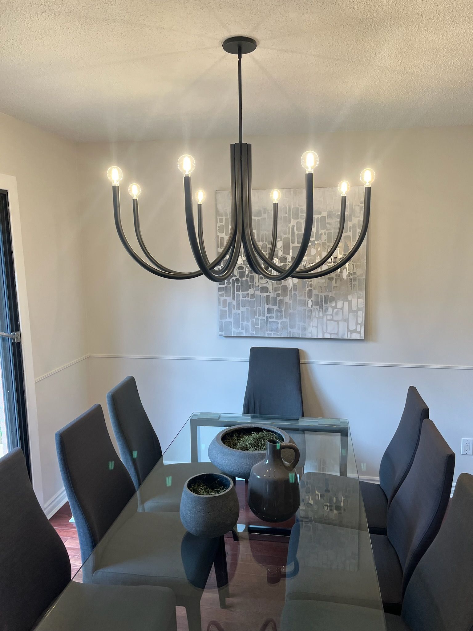 Dining room with a chandelier above a glass table and gray chairs. A piece of art hangs on the wall.