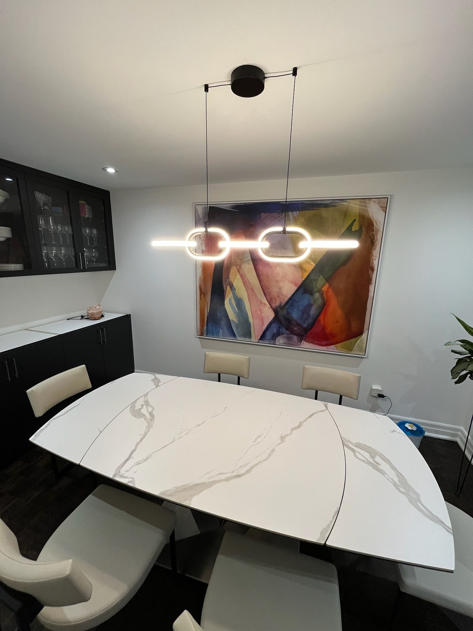 Dining room with modern light fixture, white table, chairs, and colorful artwork.