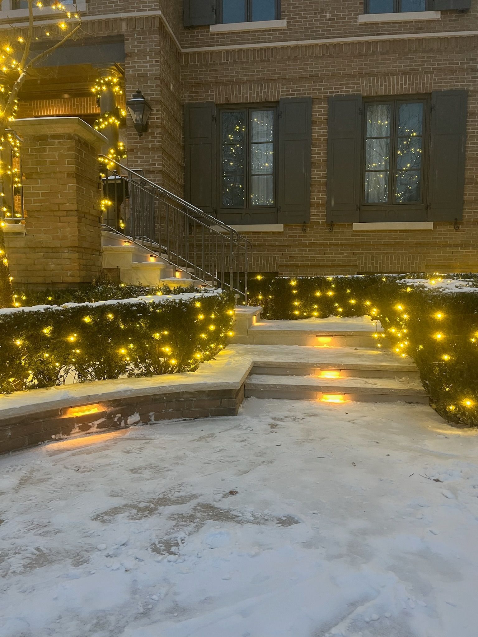 Snowy house exterior, lit with golden Christmas lights on bushes, stairs, and windows.