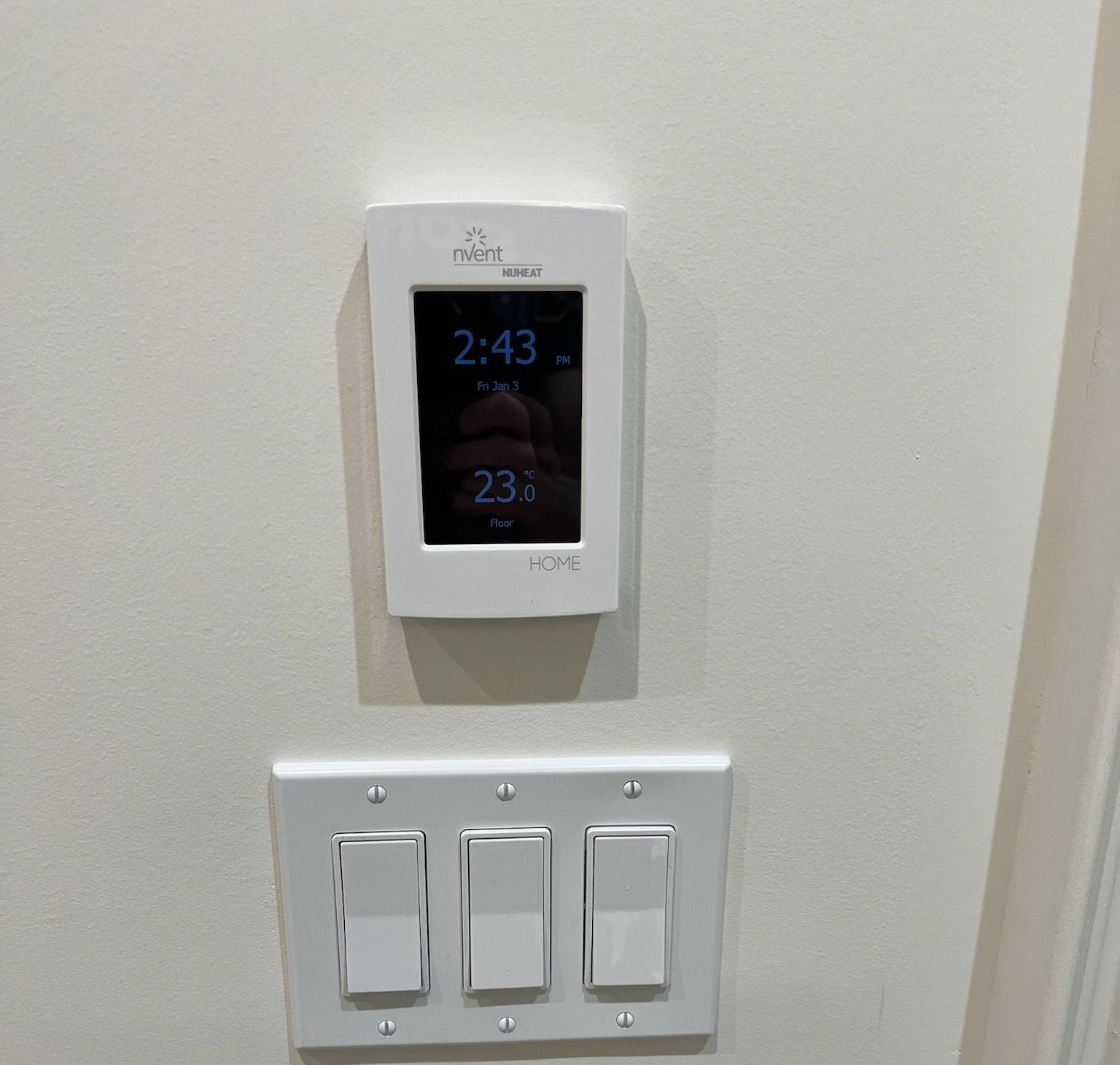 Thermostat and light switches on a beige wall, time displays as 2:43 and 23 degrees.