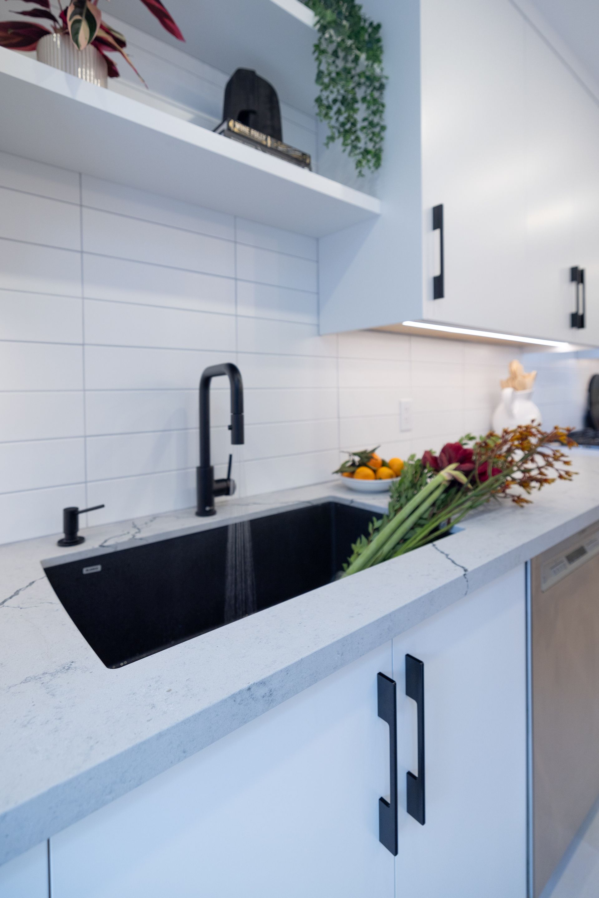 Modern kitchen sink with black faucet, soap dispenser, and produce. White cabinets and tile, gray countertop.