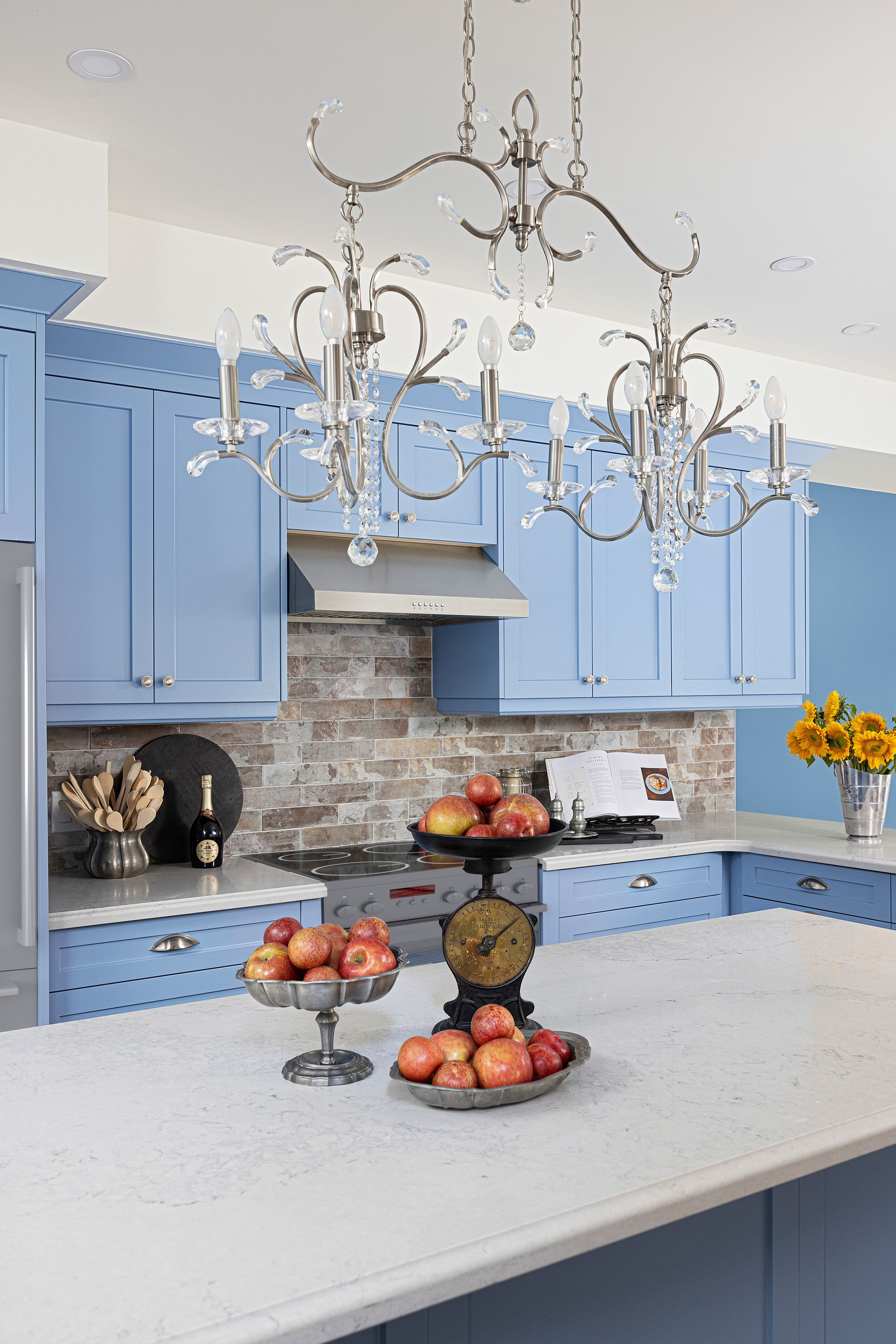 Blue kitchen with white countertops and crystal chandeliers; fruit displayed on a scale and in bowls.