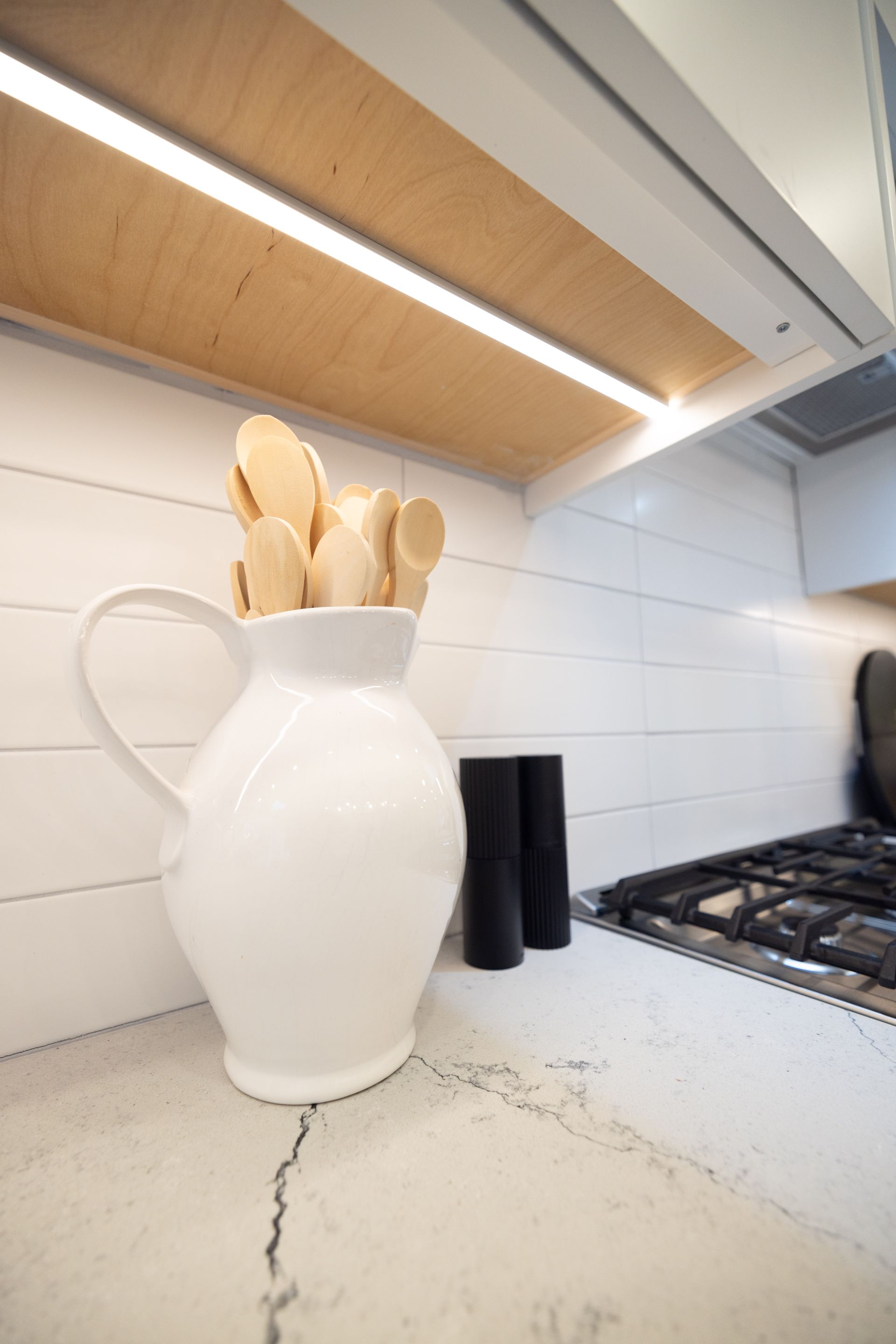 White pitcher with wooden spoons, near stove on white countertop, underlit wooden shelf.