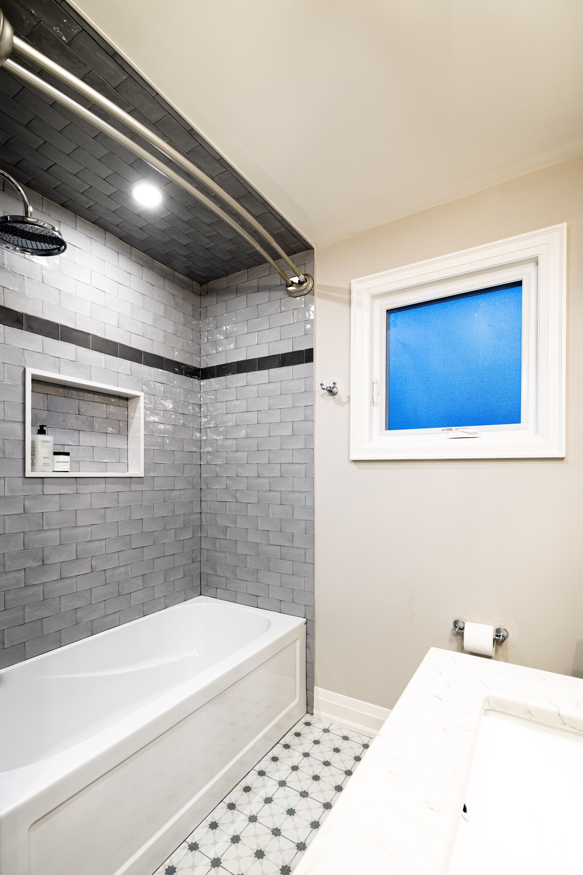 Bathroom with a white tub, gray tiled shower, and a small blue window.