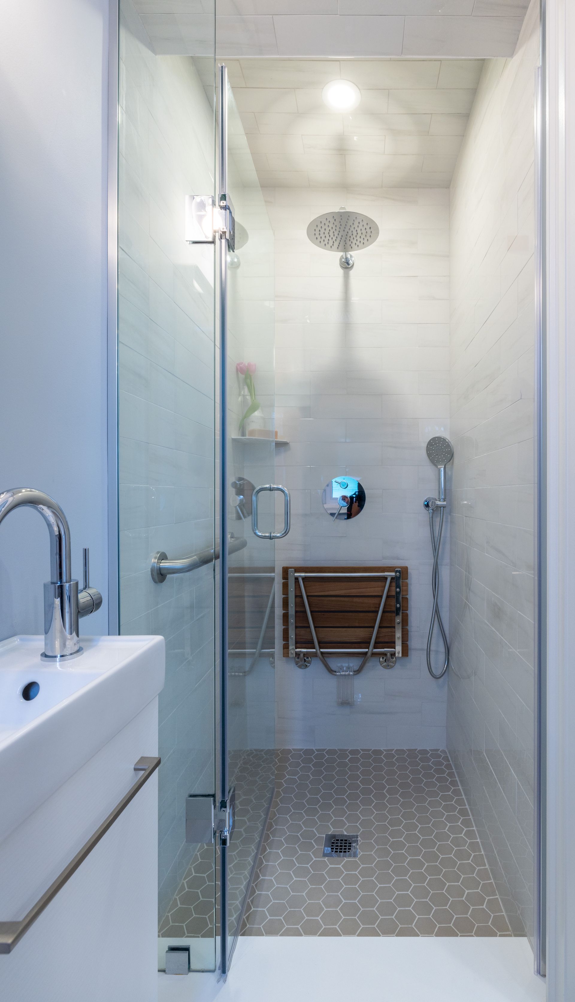 Small, white-tiled shower with a glass door, handrail, and fold-down wooden seat.