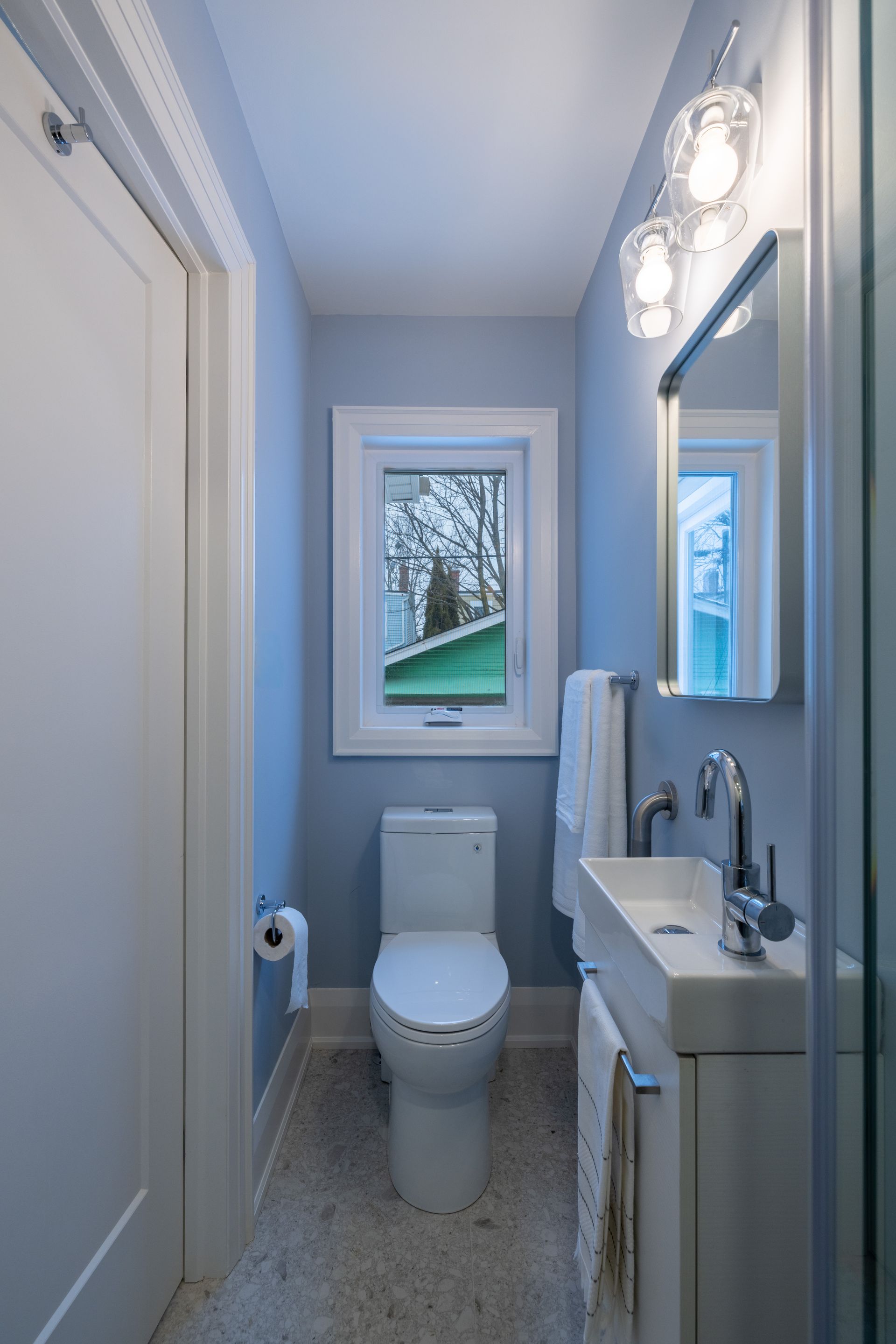 Narrow bathroom with light blue walls, white toilet, vanity, and window.