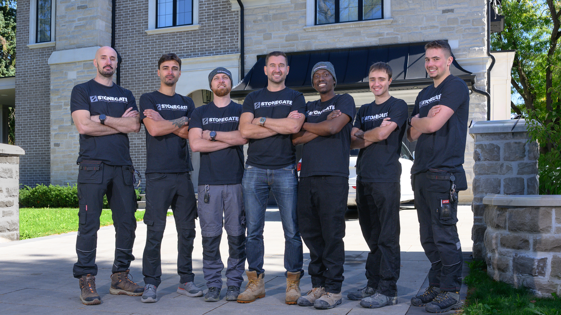 Seven people in black t-shirts and work clothes stand with arms crossed in front of a stone building.