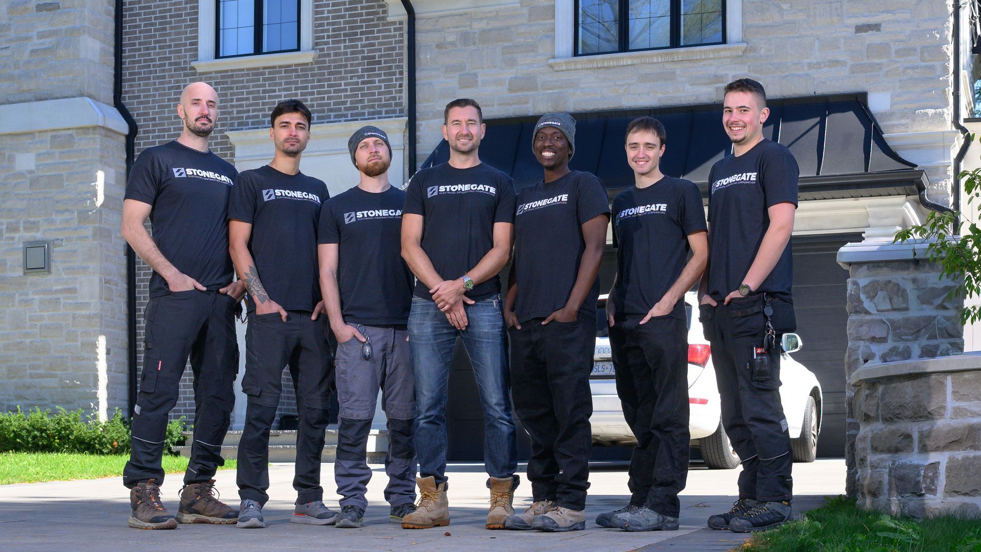 Seven people wearing black shirts pose in front of a stone building with a garage door.