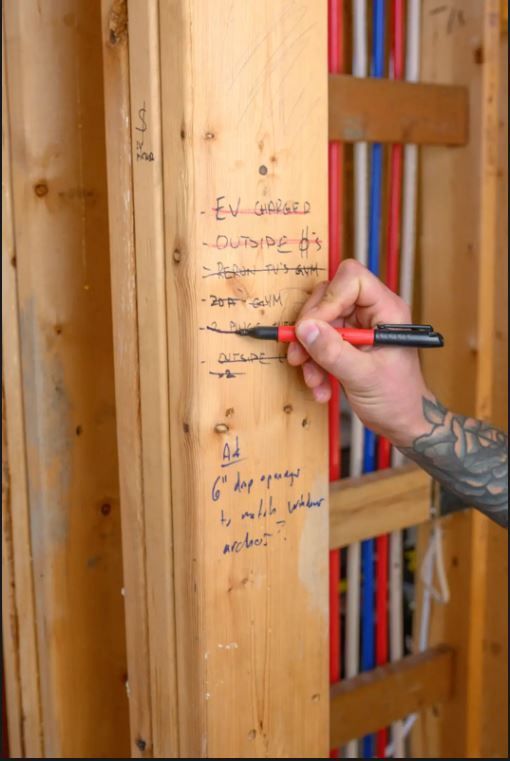 Hand writing notes on a wooden stud next to colored plumbing pipes in a construction setting.