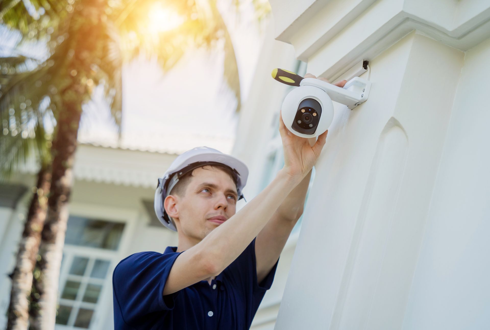 Man in hard hat installs a security camera on a white building exterior under sunlight.