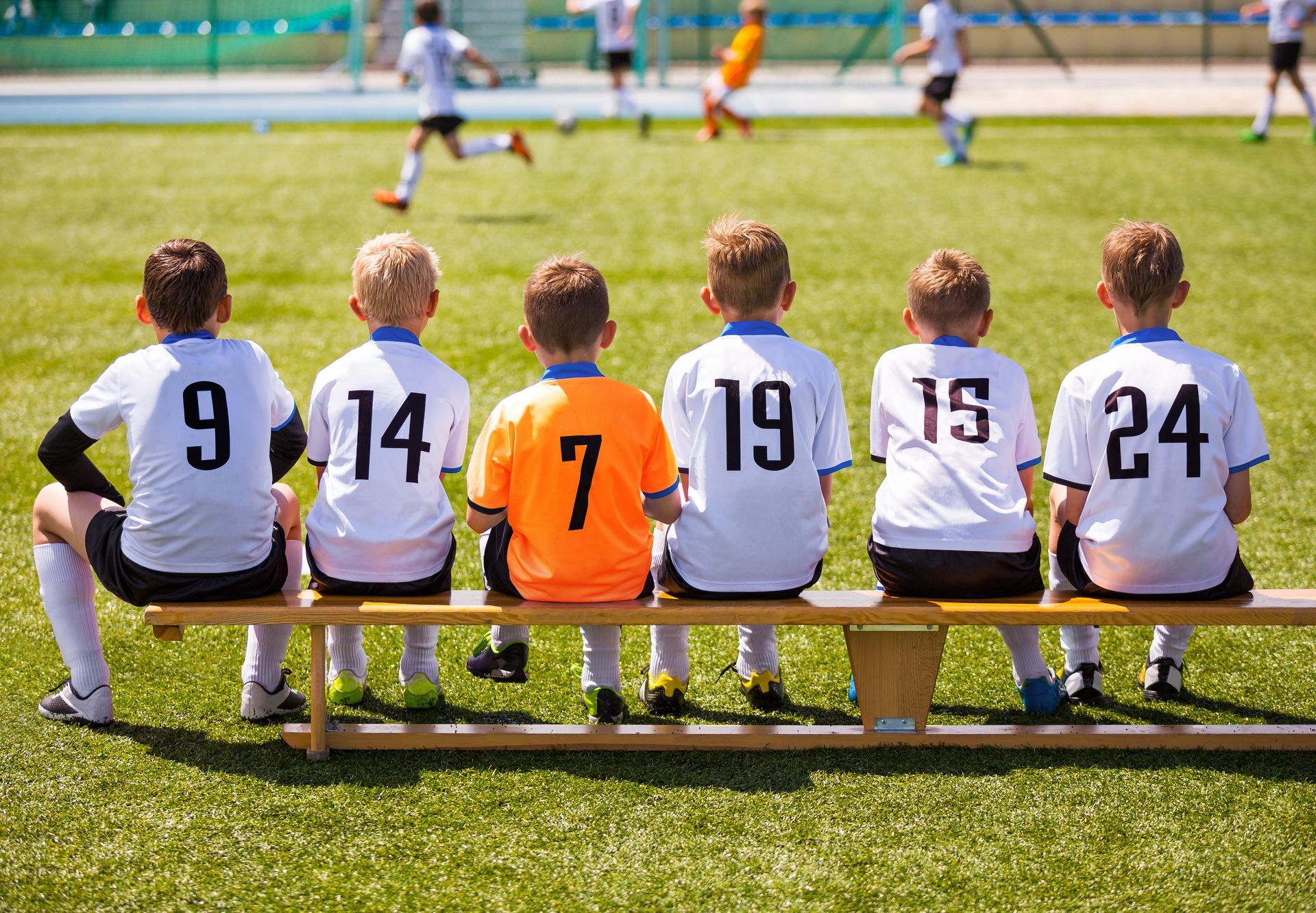 Six young soccer players sit on a bench, watching a game. Players wear white or orange jerseys with numbers on their backs.