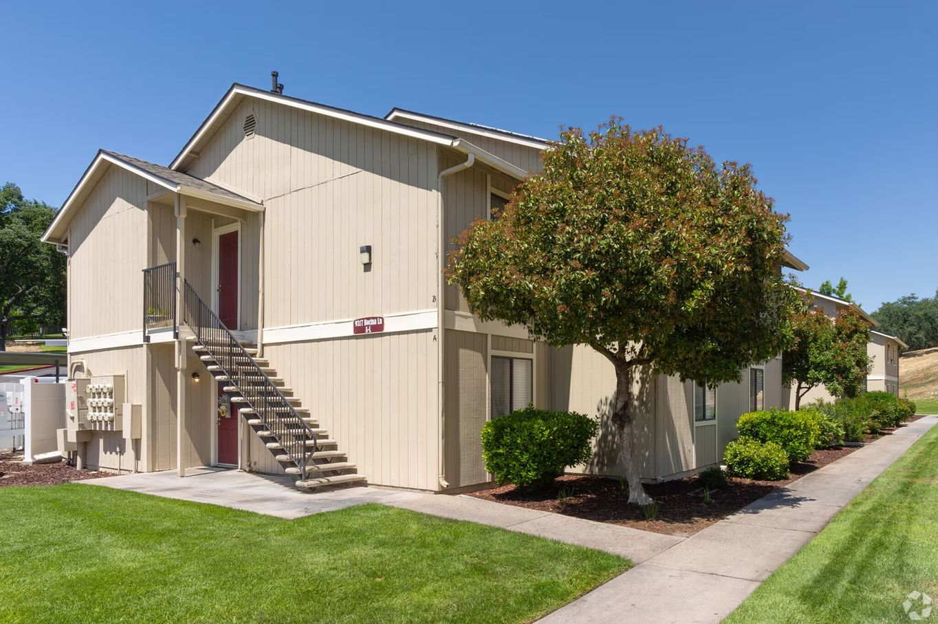 Two-story apartment building with beige siding, dark staircases, and a green lawn on a sunny day.