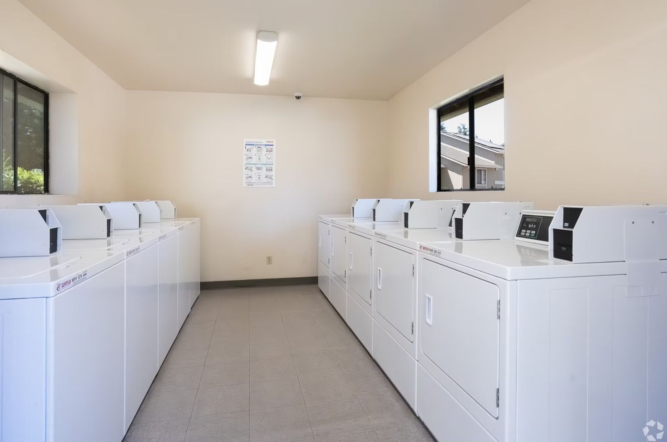 Laundromat with rows of white washing machines and dryers, light walls, and small windows.