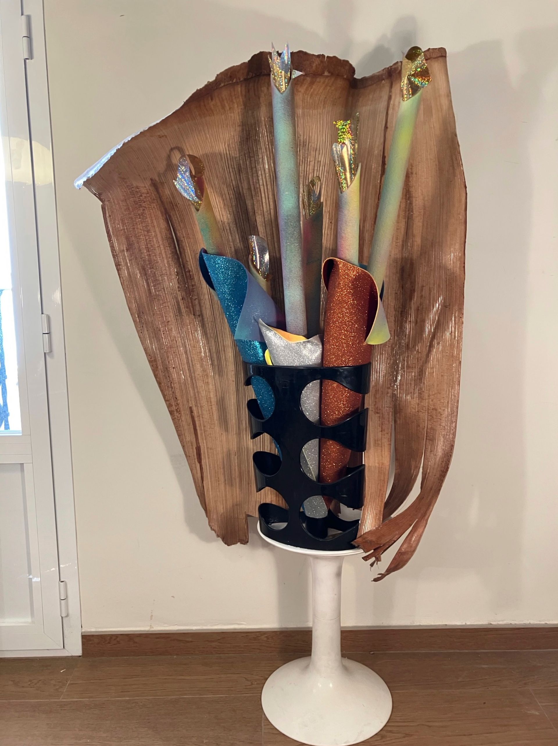 Skulpturales Kunstwerk: Palmenblatt mit farbigen Zylindern, auf einem weißen Sockel.