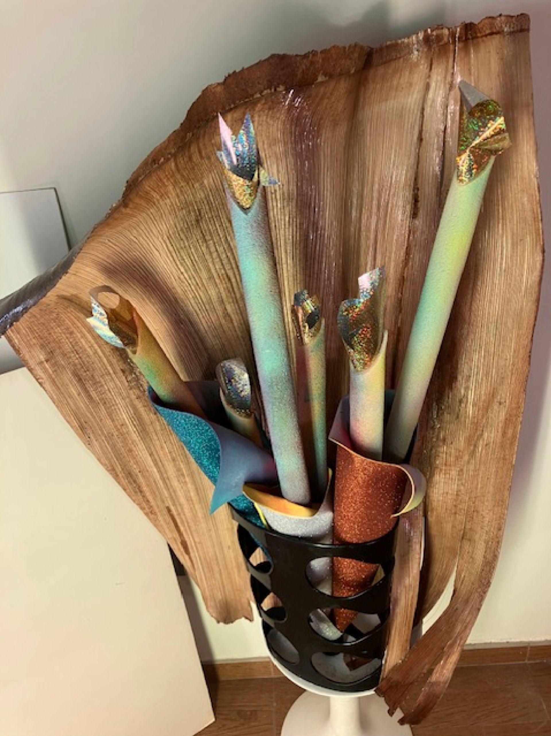 Dekoratives Kunstwerk: bunte Röhren in einem schwarz-weißen Ausschnitthalter vor einem braunen, strukturierten Hintergrund.