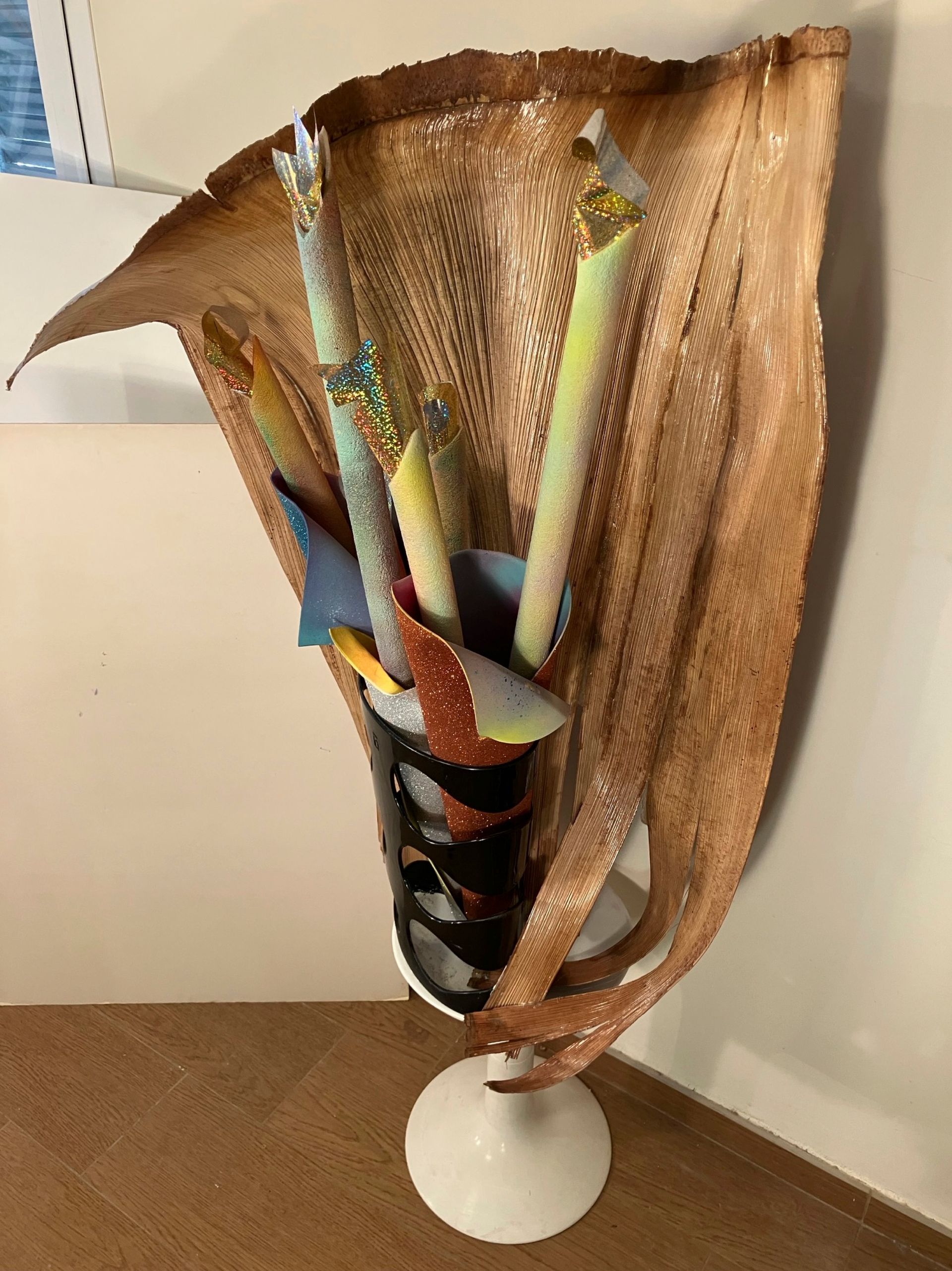 Skulpturale Vase mit getrockneten Blättern und bunten Stäben, stehend auf einem weißen Sockel vor einer beigen Wand.