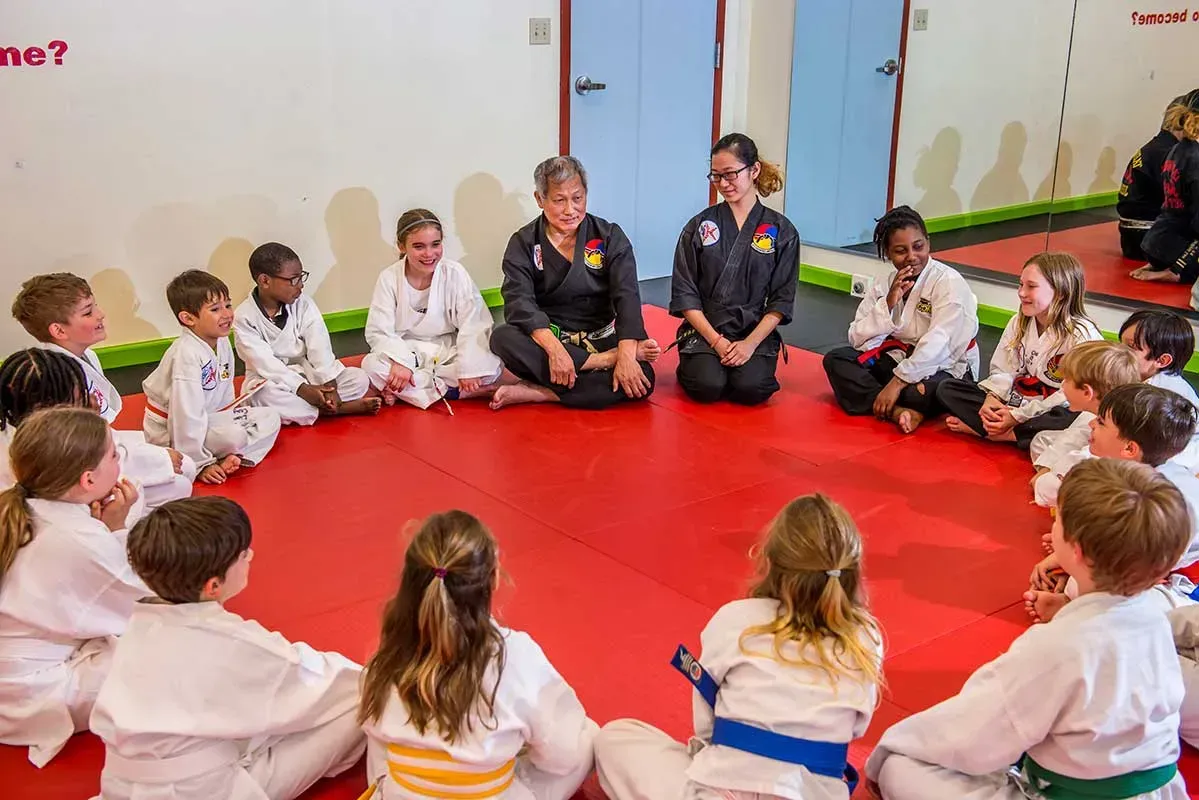 Children in martial arts uniforms sit in a circle with instructors on a red mat inside a gym