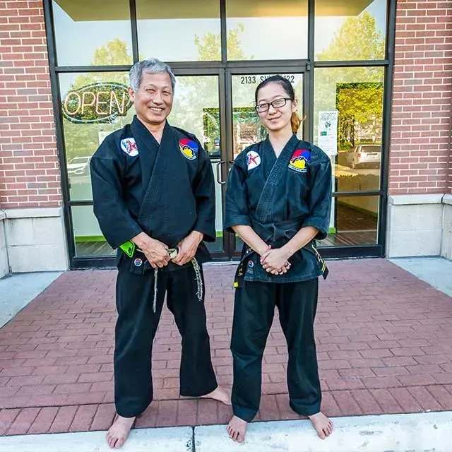 Two people in black martial arts uniforms stand outside a storefront.