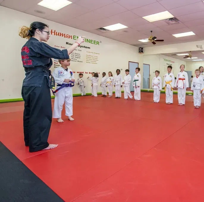 Martial arts instructor in black uniform instructs children in white gi, on a red mat