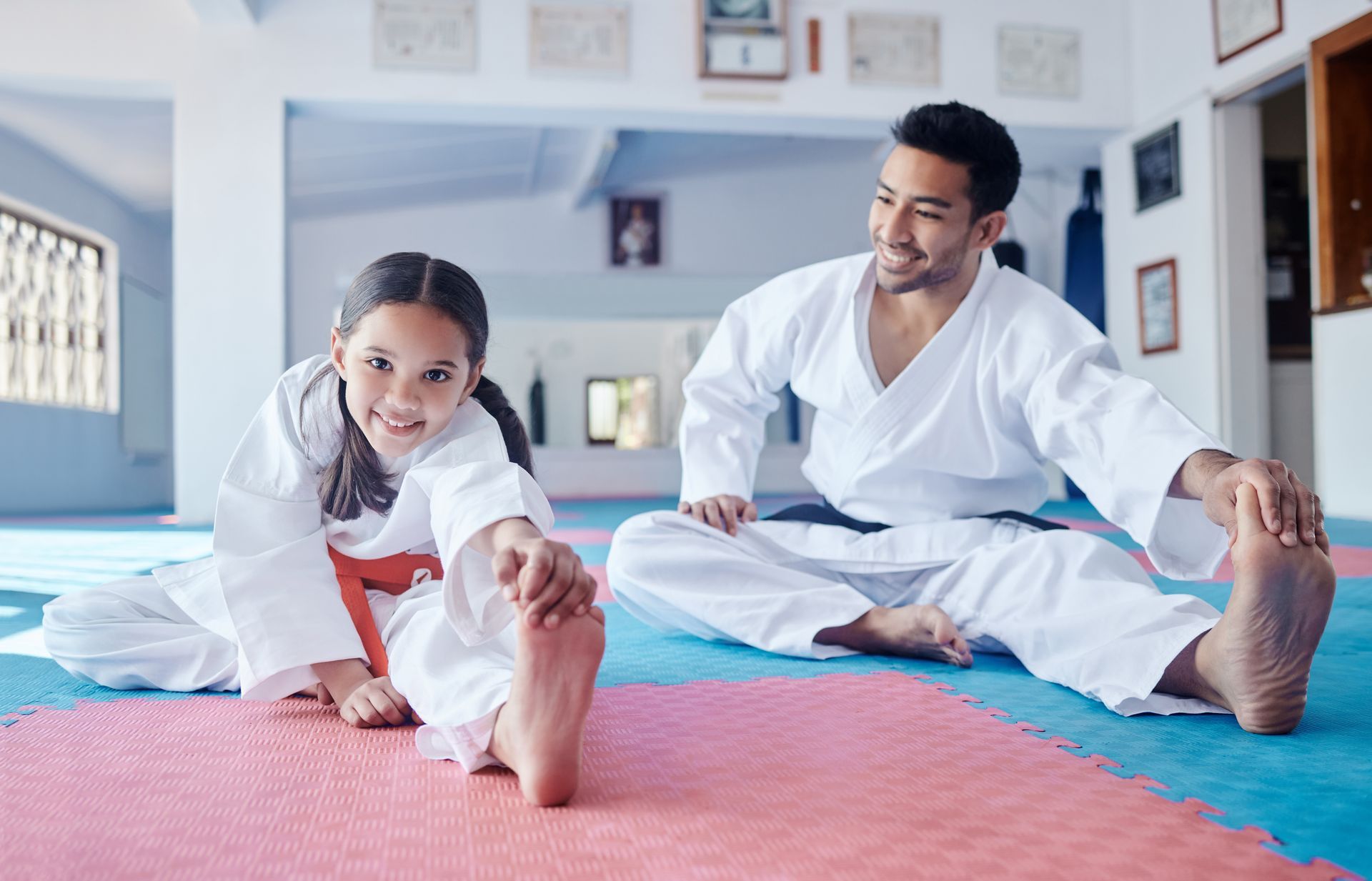 Man and child in white karate uniforms stretching on mats in a dojo.