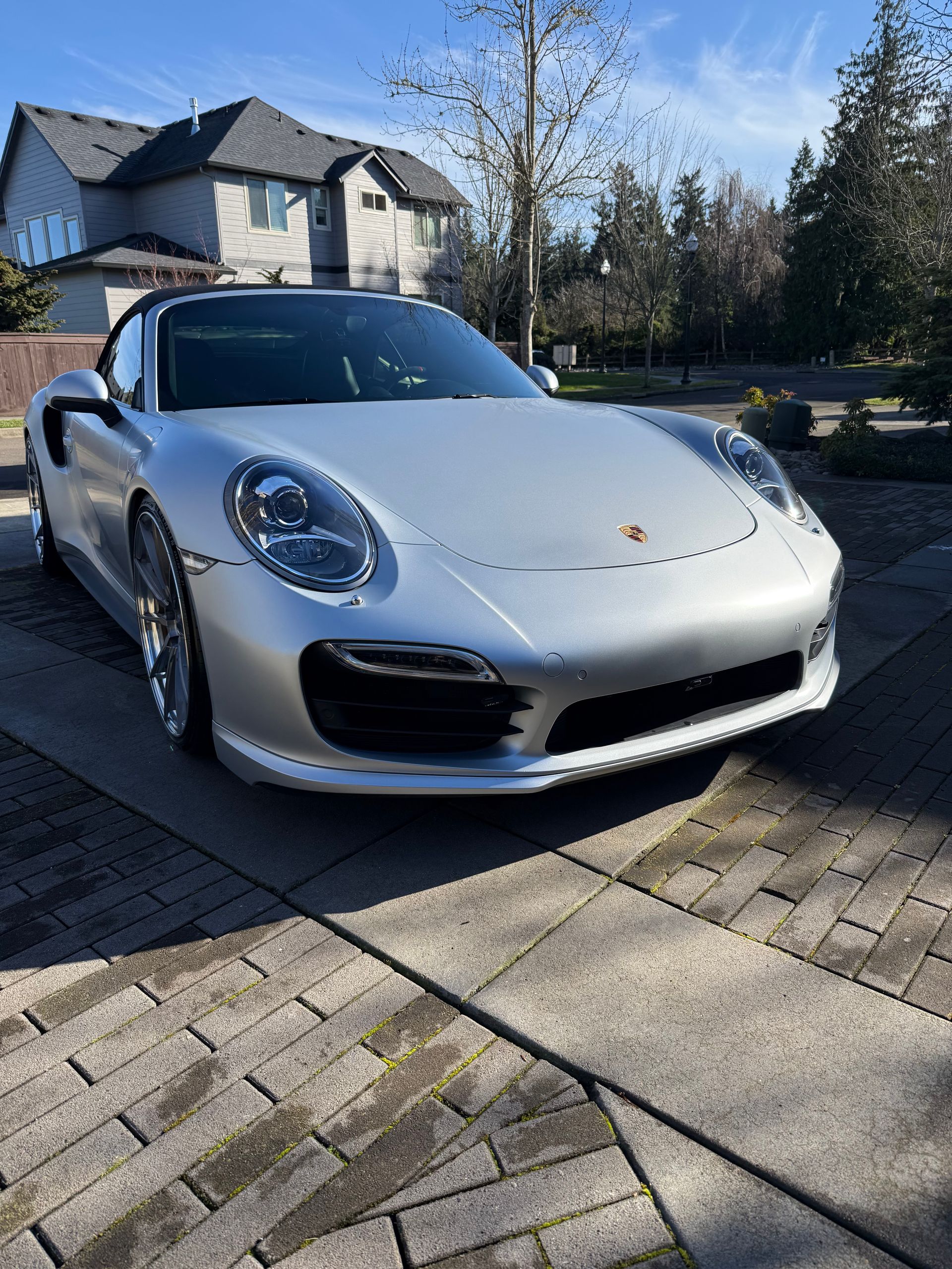 A silver Porsche convertible parked on a paved driveway in front of a residential house under a clear blue sky.