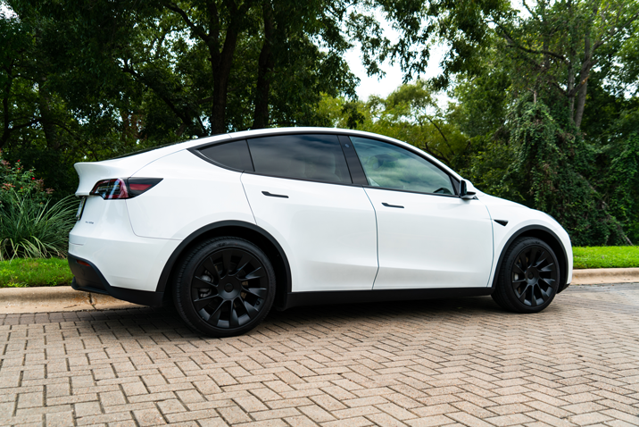 A white Tesla Model Y with black wheels parked on a paved driveway surrounded by trees and green grass.