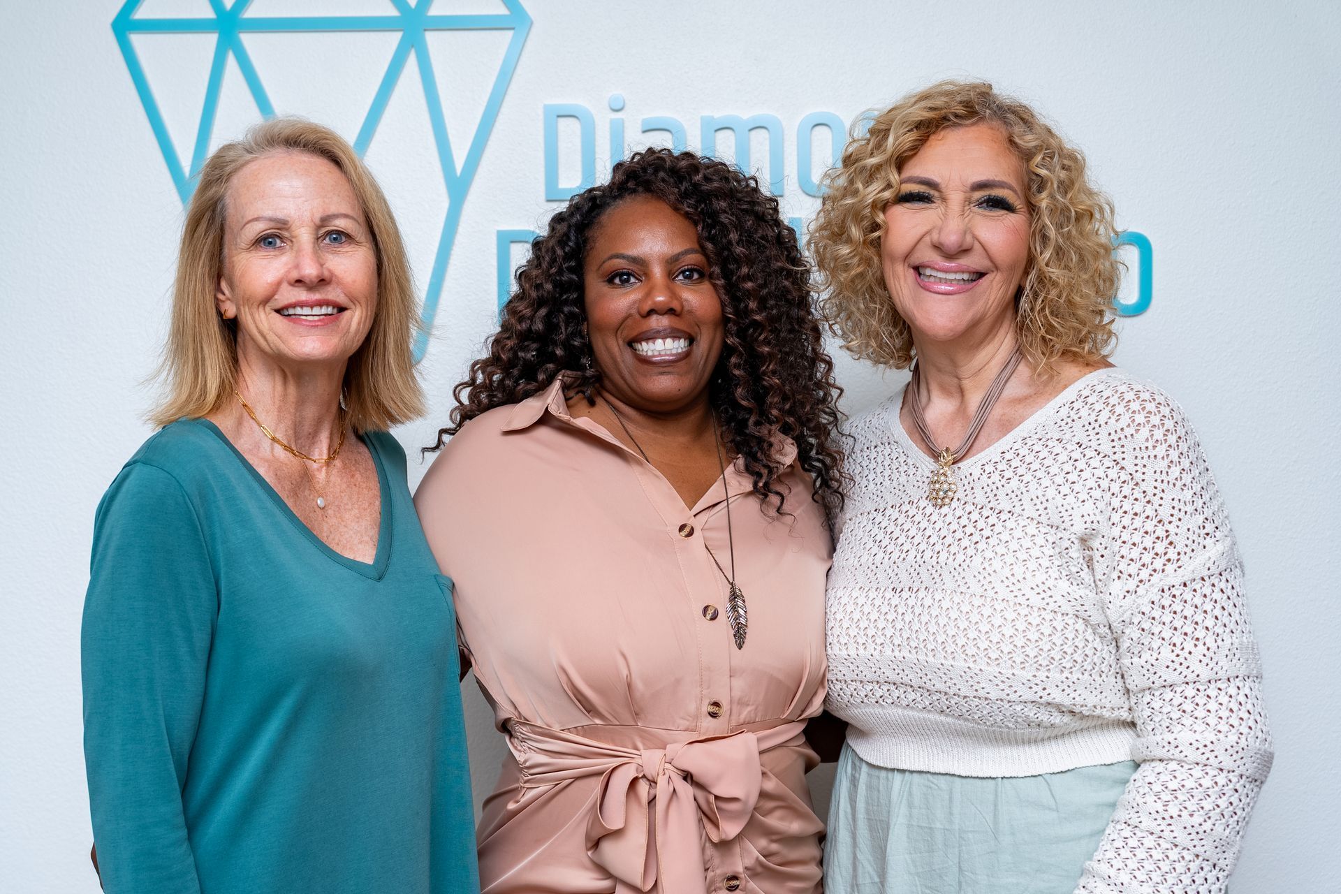 Three women posing, smiling. Diamond logo on wall.