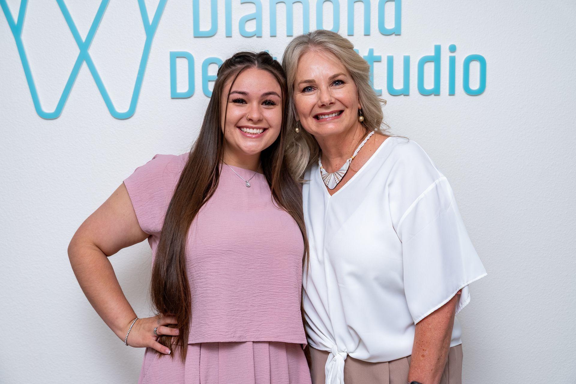 Two smiling women pose in front of a turquoise logo on a white wall.