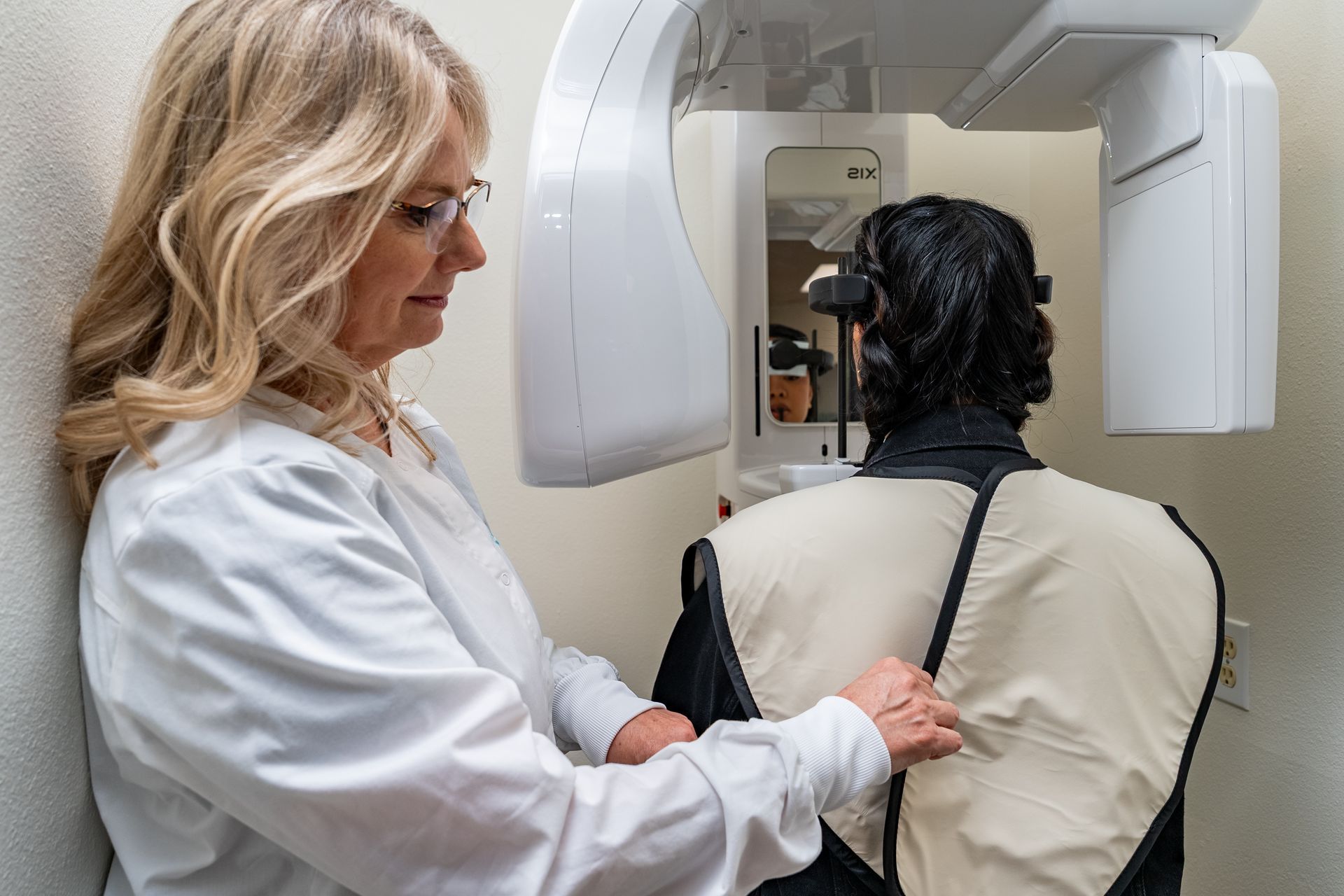 Dentist adjusting a patient's lead apron for a dental X-ray in a white-walled room.