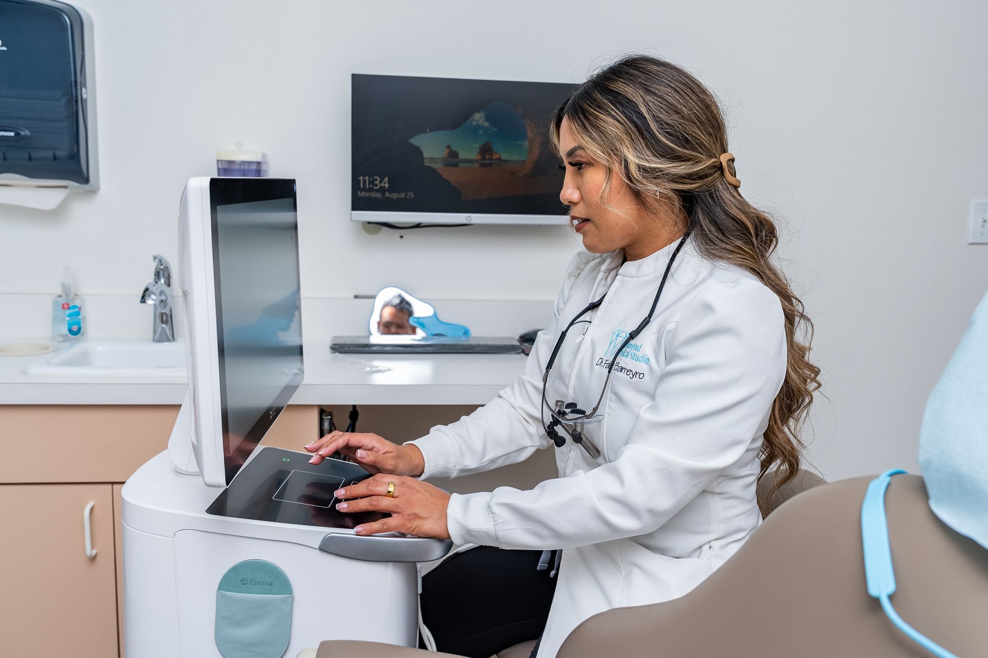 Dentist in white coat using a computer, seated in a dental office.