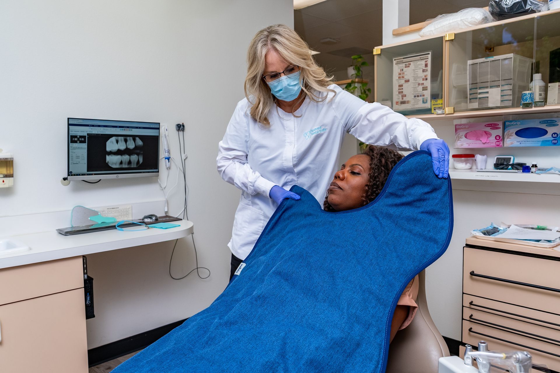 Dentist in mask and gloves draping a blue cloth over a patient in a dental chair.