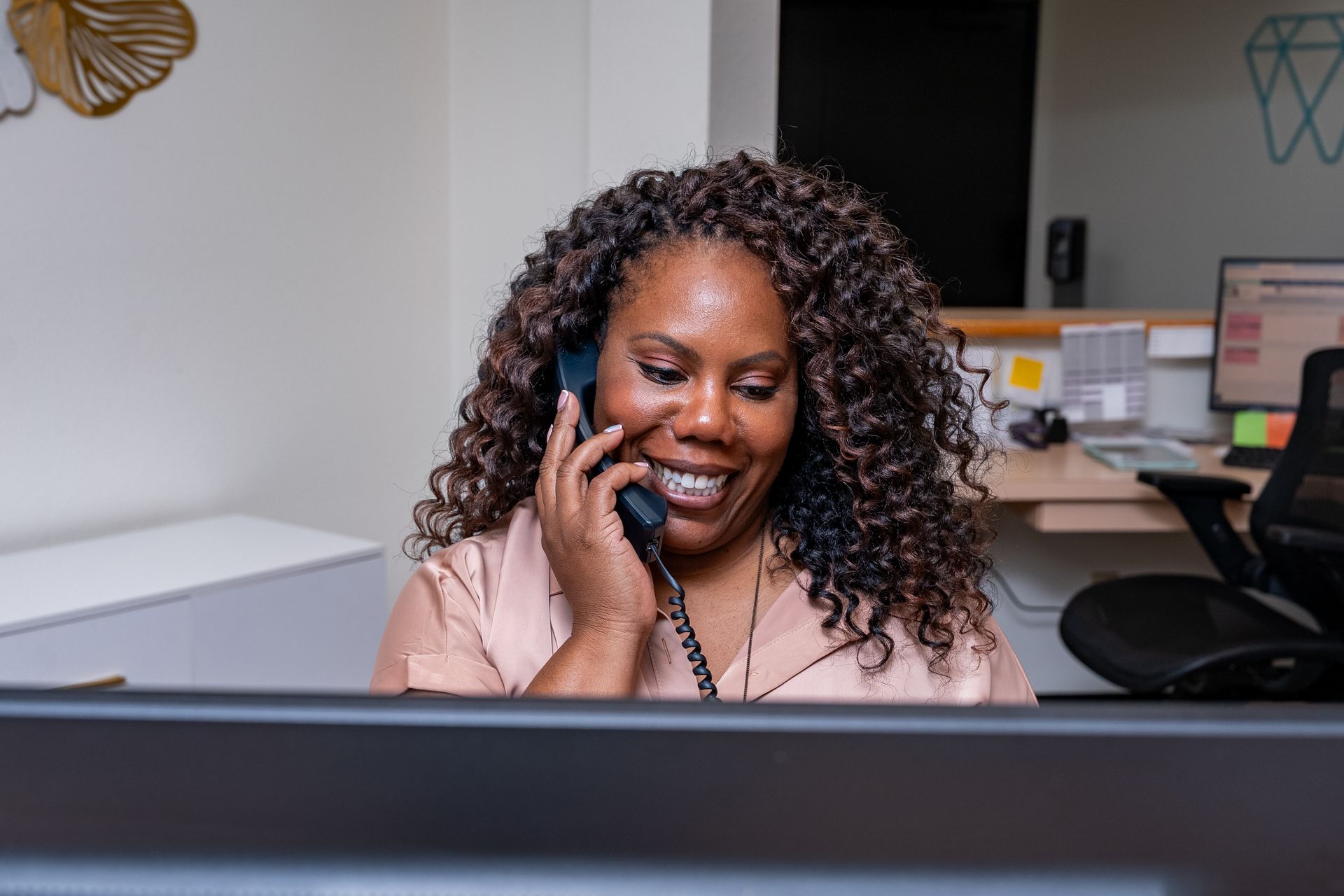 Woman with curly hair smiles while talking on a phone at an office desk.