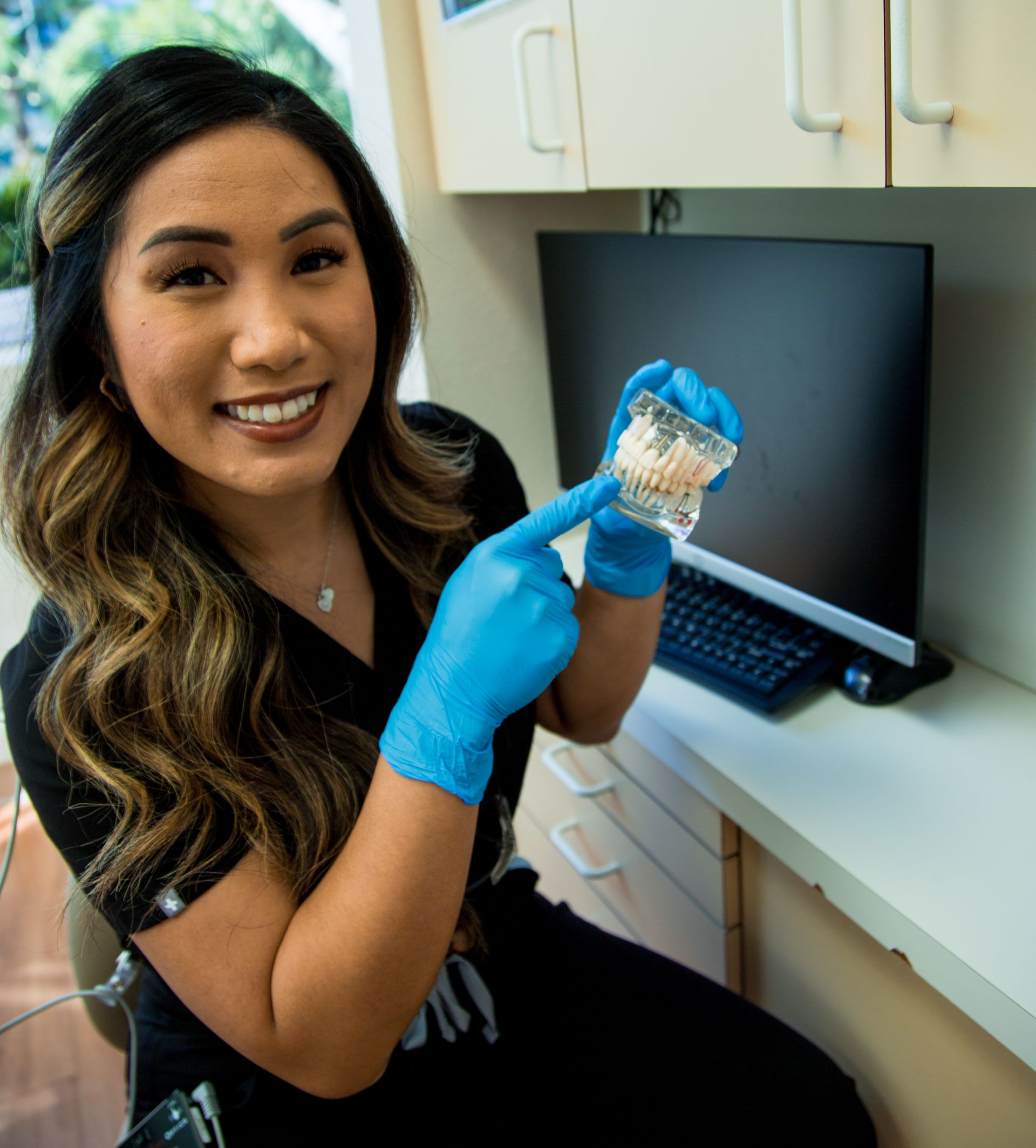 Dentist pointing to teeth model, wearing blue gloves, smiling in a dental office.