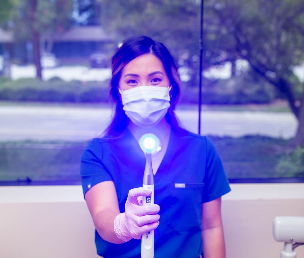 Dentist in blue scrubs and mask holding a blue light, looking at the camera.