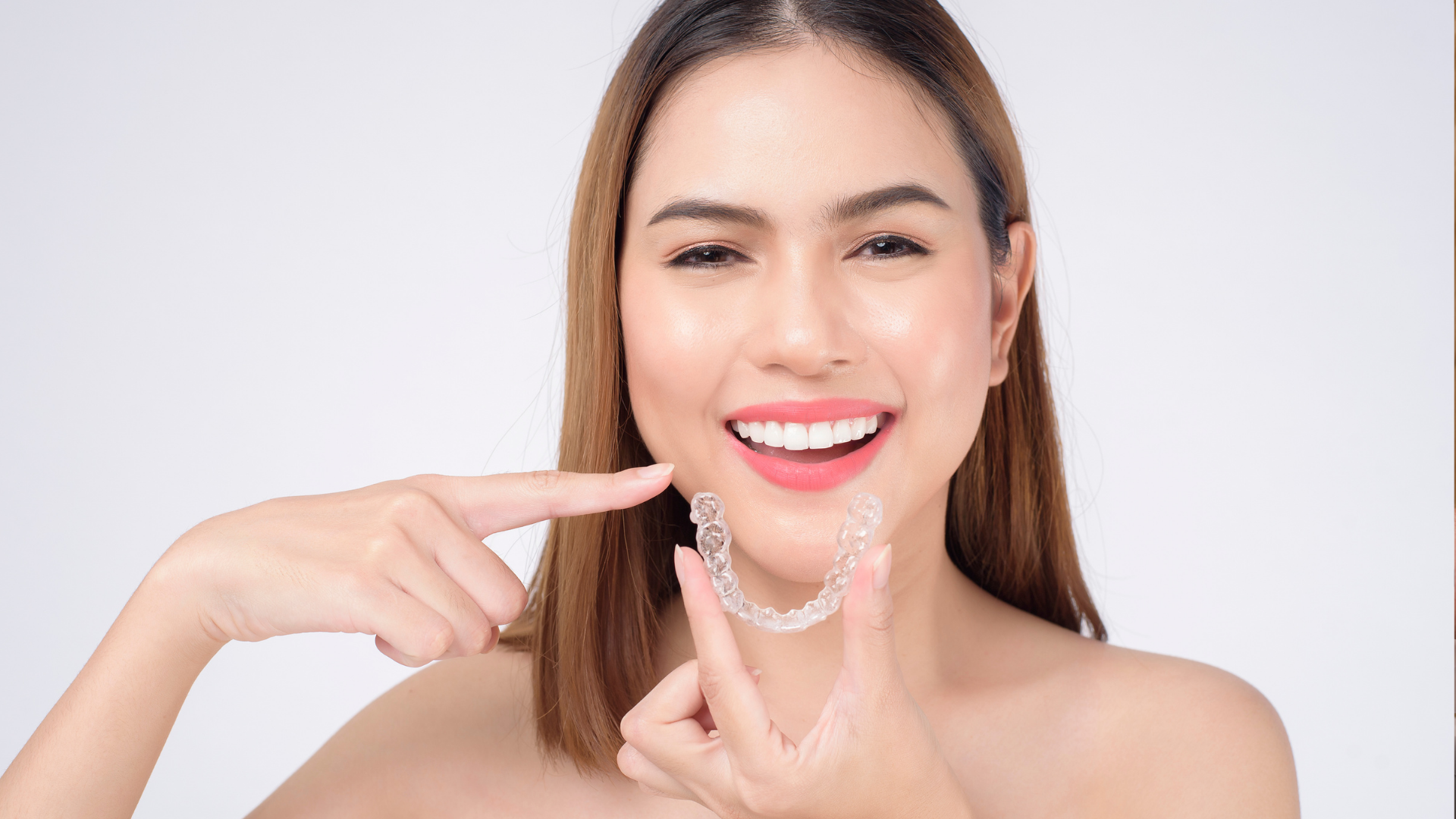 Woman smiling, holding clear aligners, pointing to teeth, against a white background.