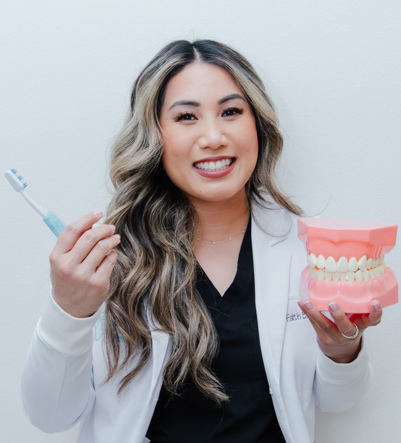 Dentist holding toothbrush and dental model, smiling. Wearing a lab coat and black scrubs.
