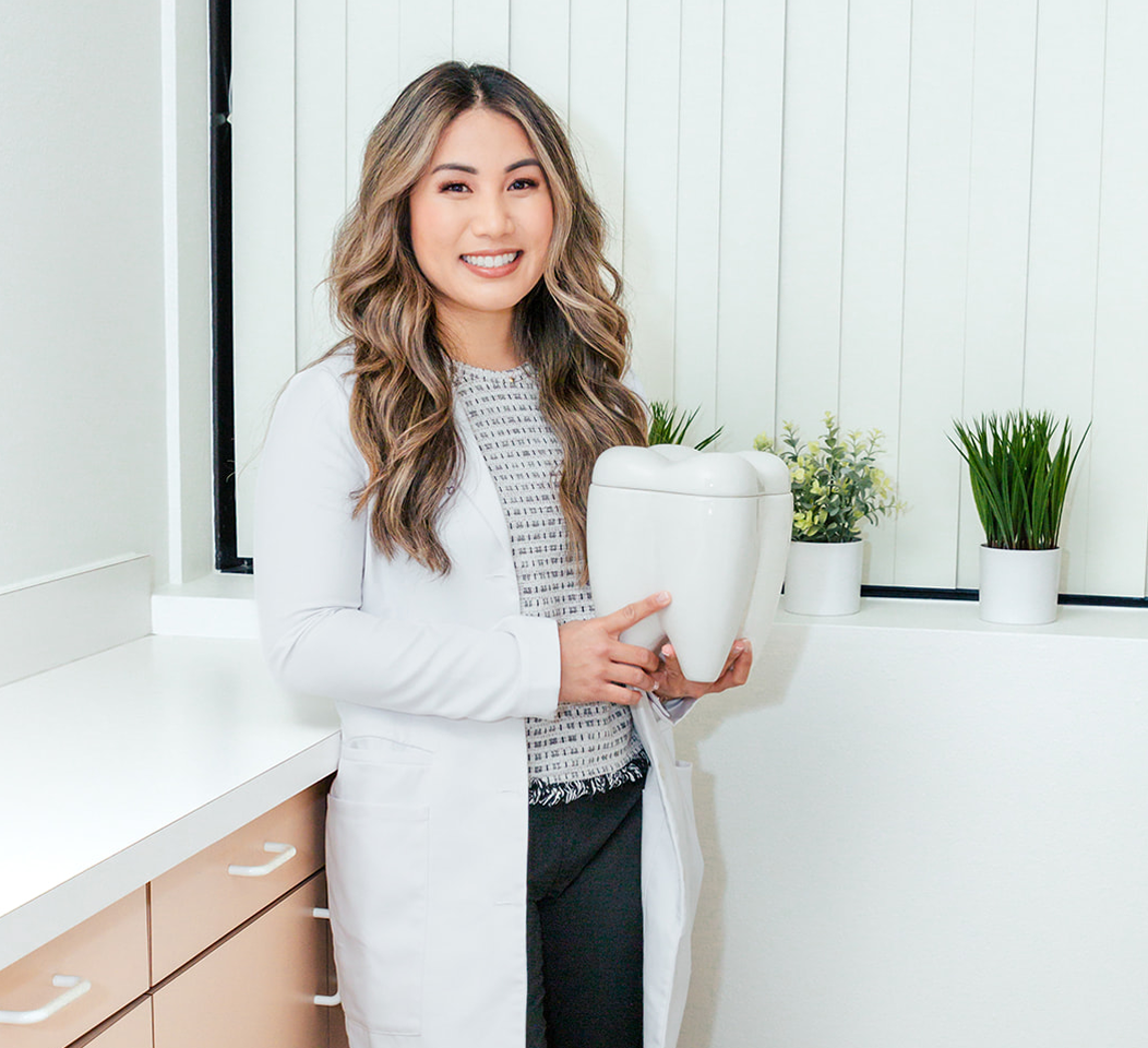 Woman dentist in lab coat holding a giant tooth, smiling in an office setting.