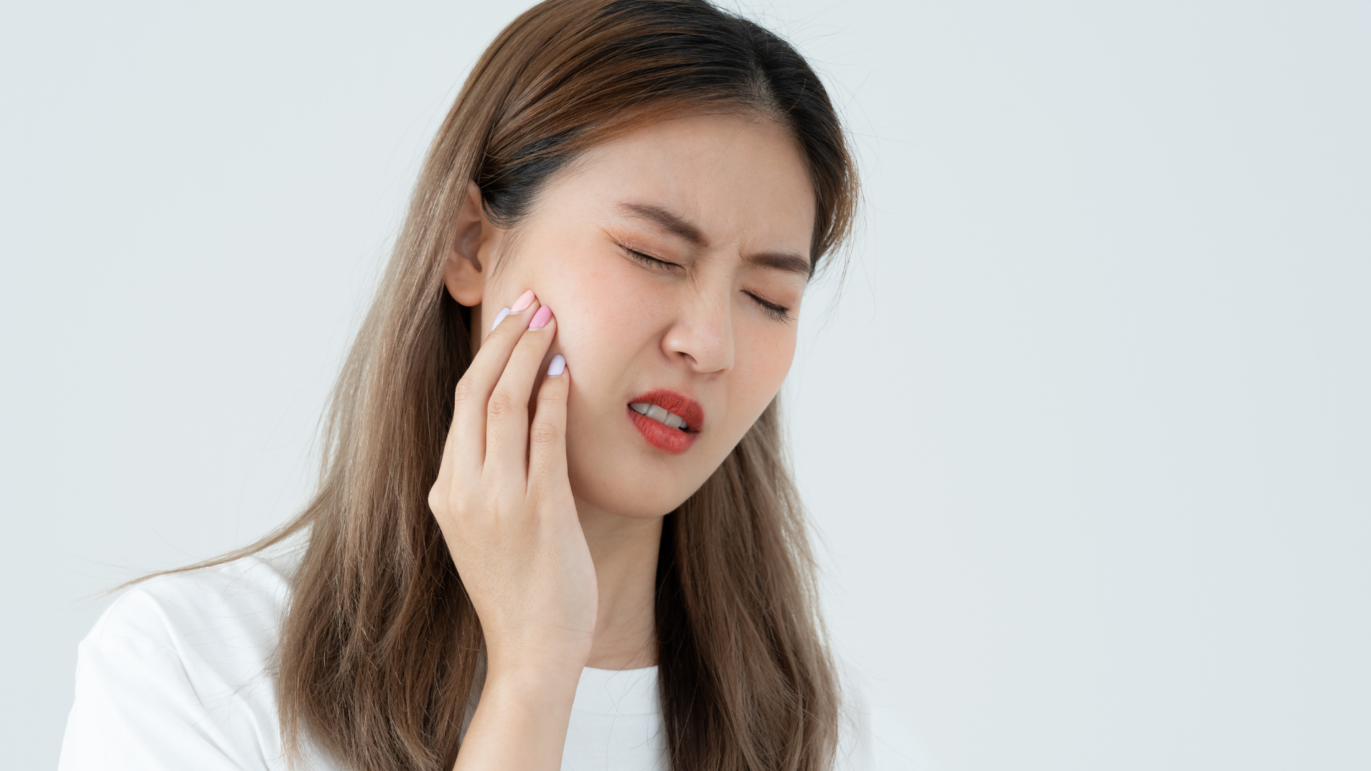 Woman in white shirt with hand on jaw, showing pain, eyes closed.