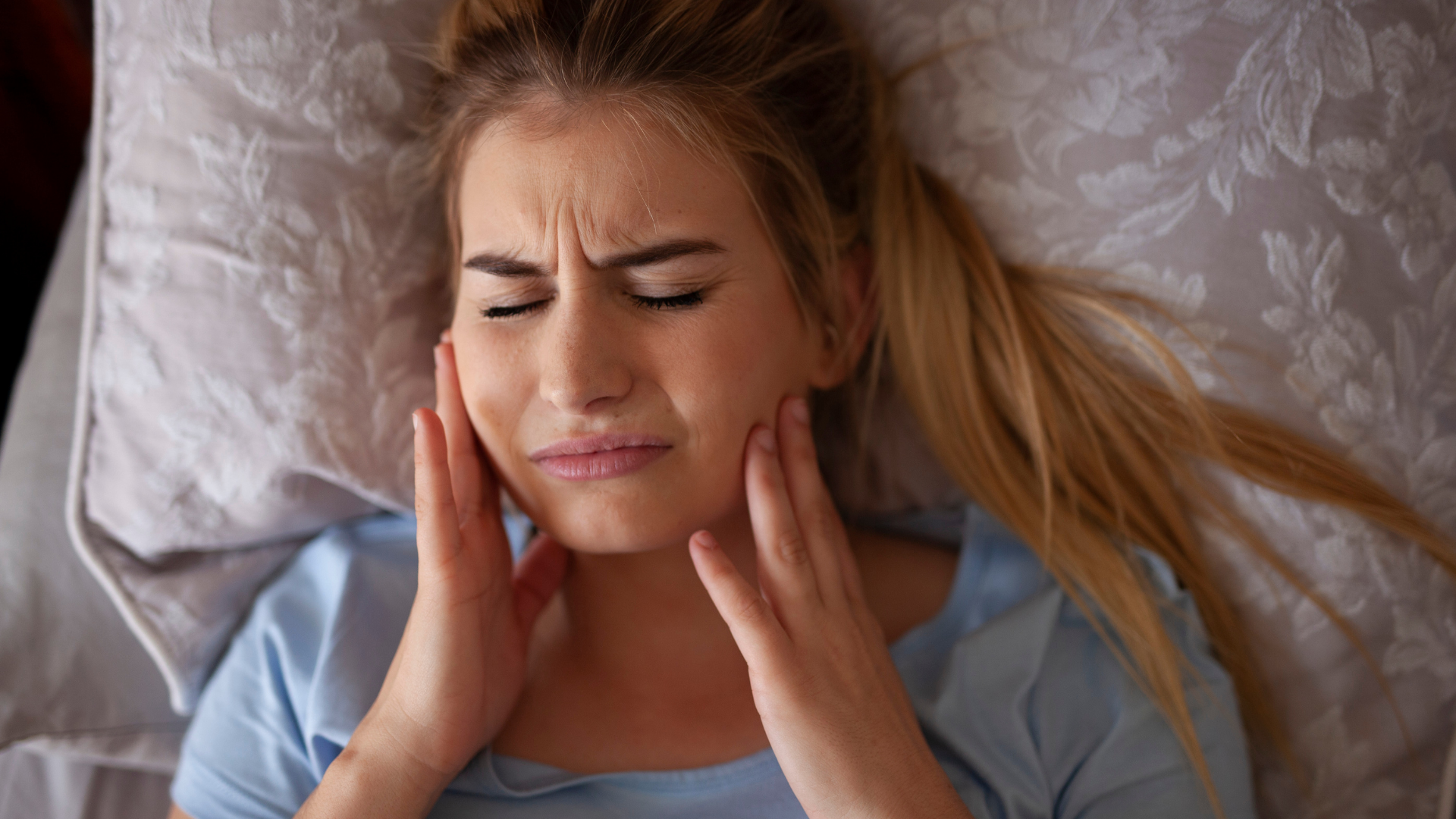 Woman lying in bed, holding cheeks, facial expression showing pain.