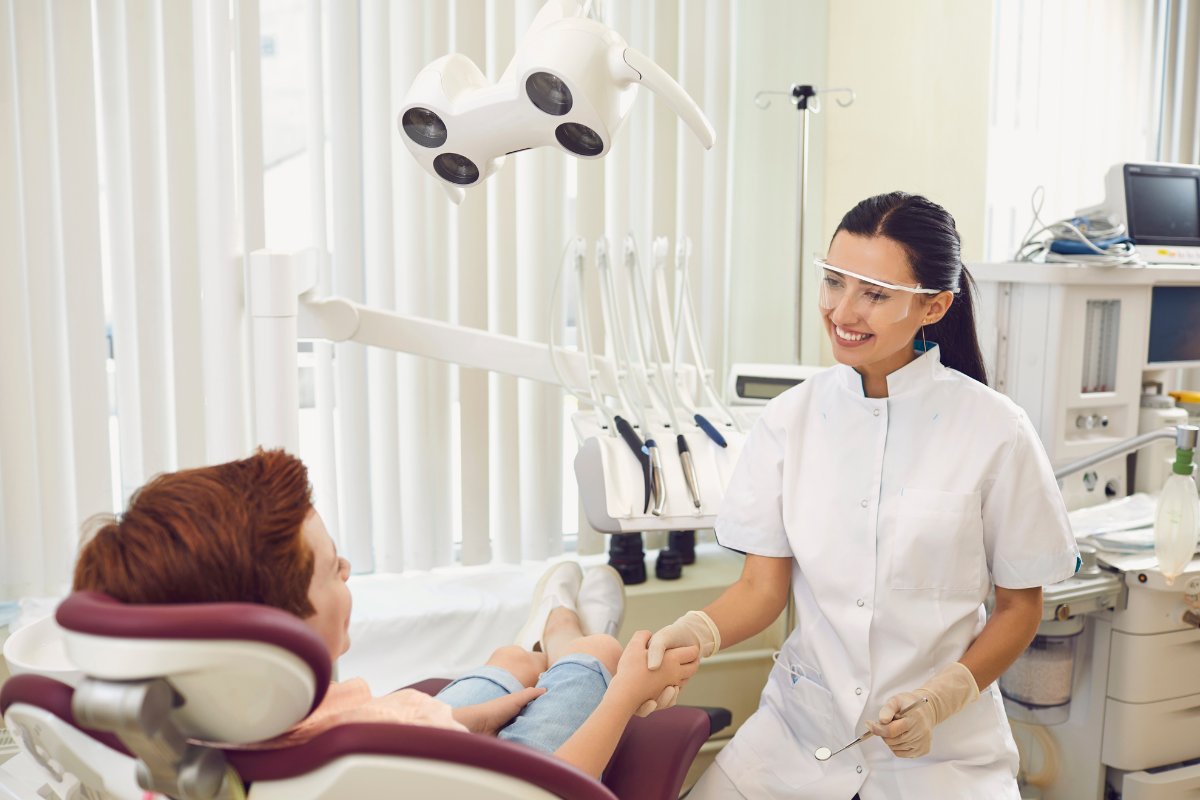 Dentist in white uniform shakes hands with patient in chair, smiling, in bright dental office.