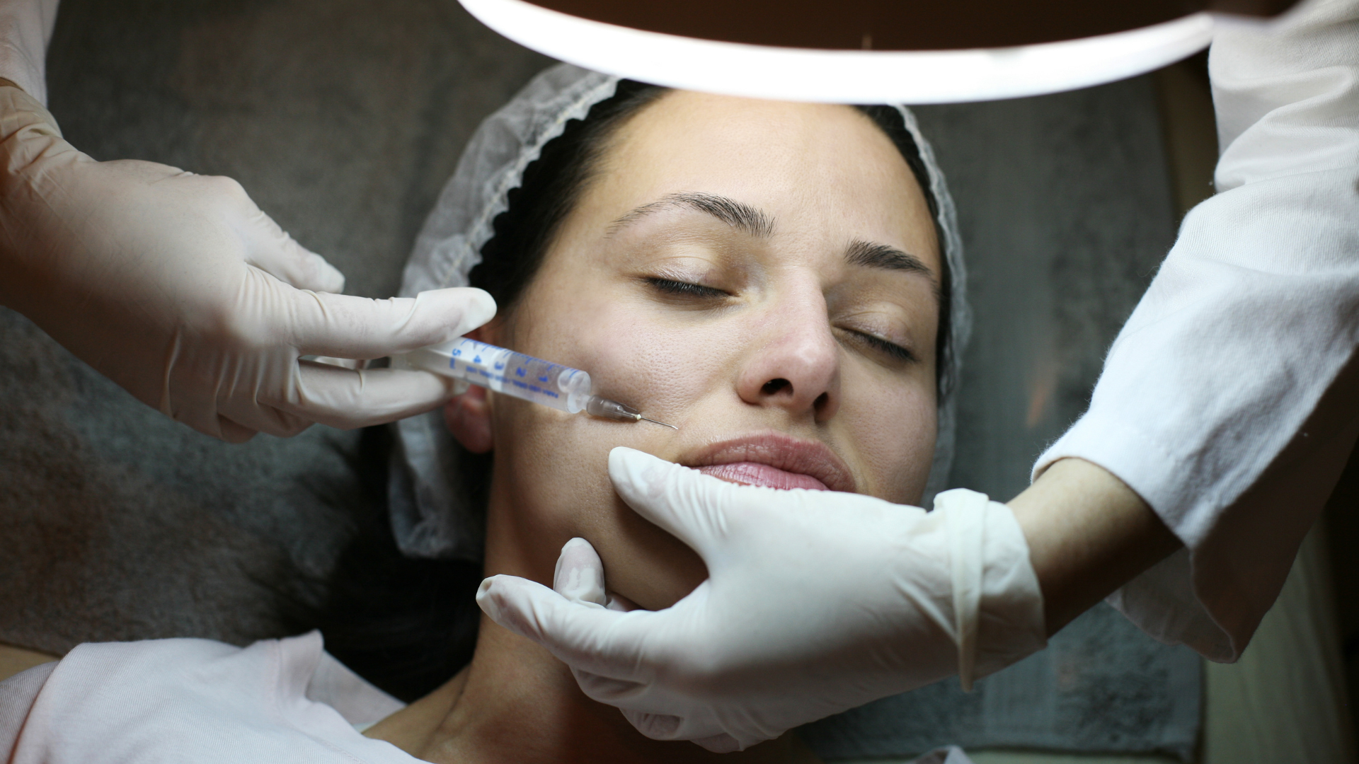Woman receiving facial injection with gloved hands, under bright light.