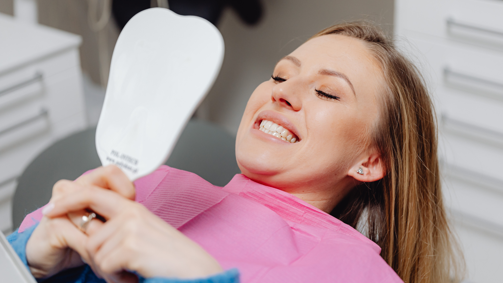 Woman in pink bib smiles while looking at her teeth in a hand mirror at a dental office.
