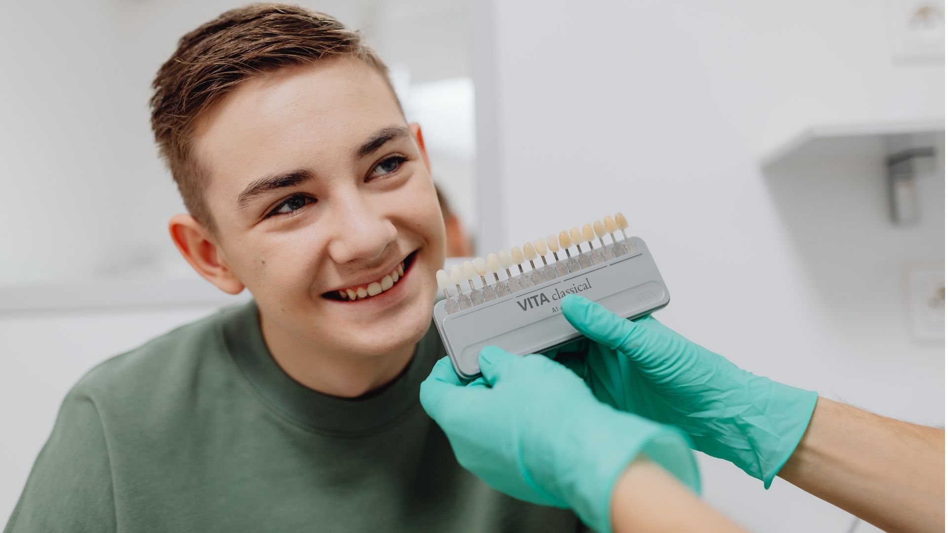 A smiling patient sits as gloved hands hold a dental shade guide near his teeth.