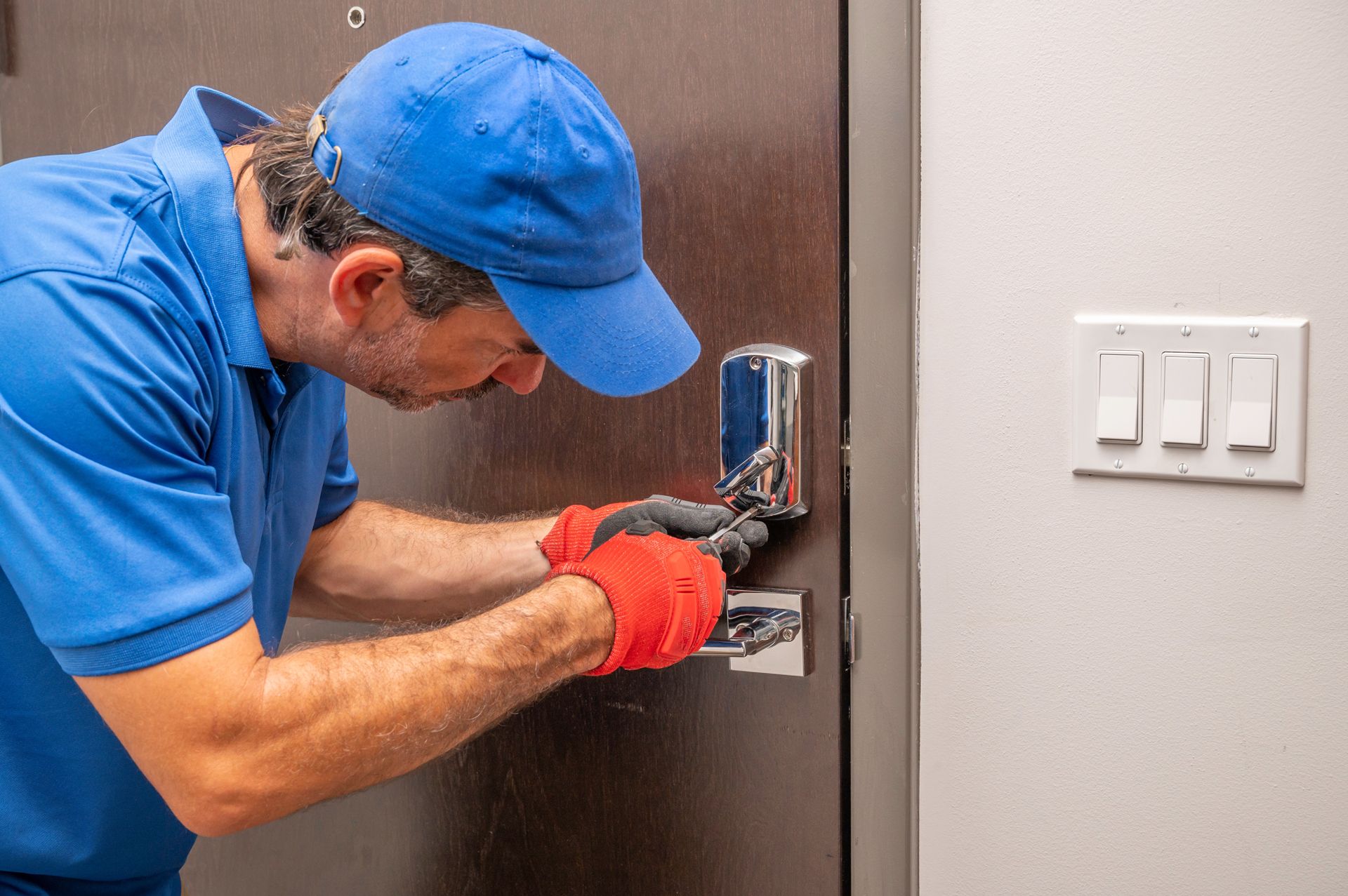 Locksmith in blue shirt and cap, red gloves, working on a door lock in a white-walled room.