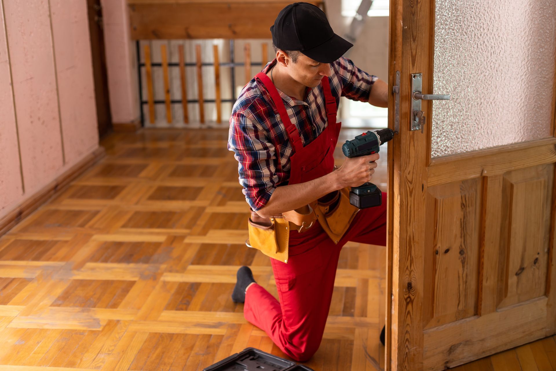 Handyman kneeling, using a drill on a wooden door in a room with wood floors.