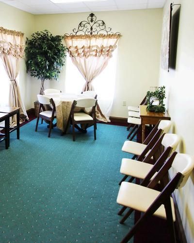 Room with a round table, chairs, curtains, and a tree, with chairs lined up on the right. Green carpet.